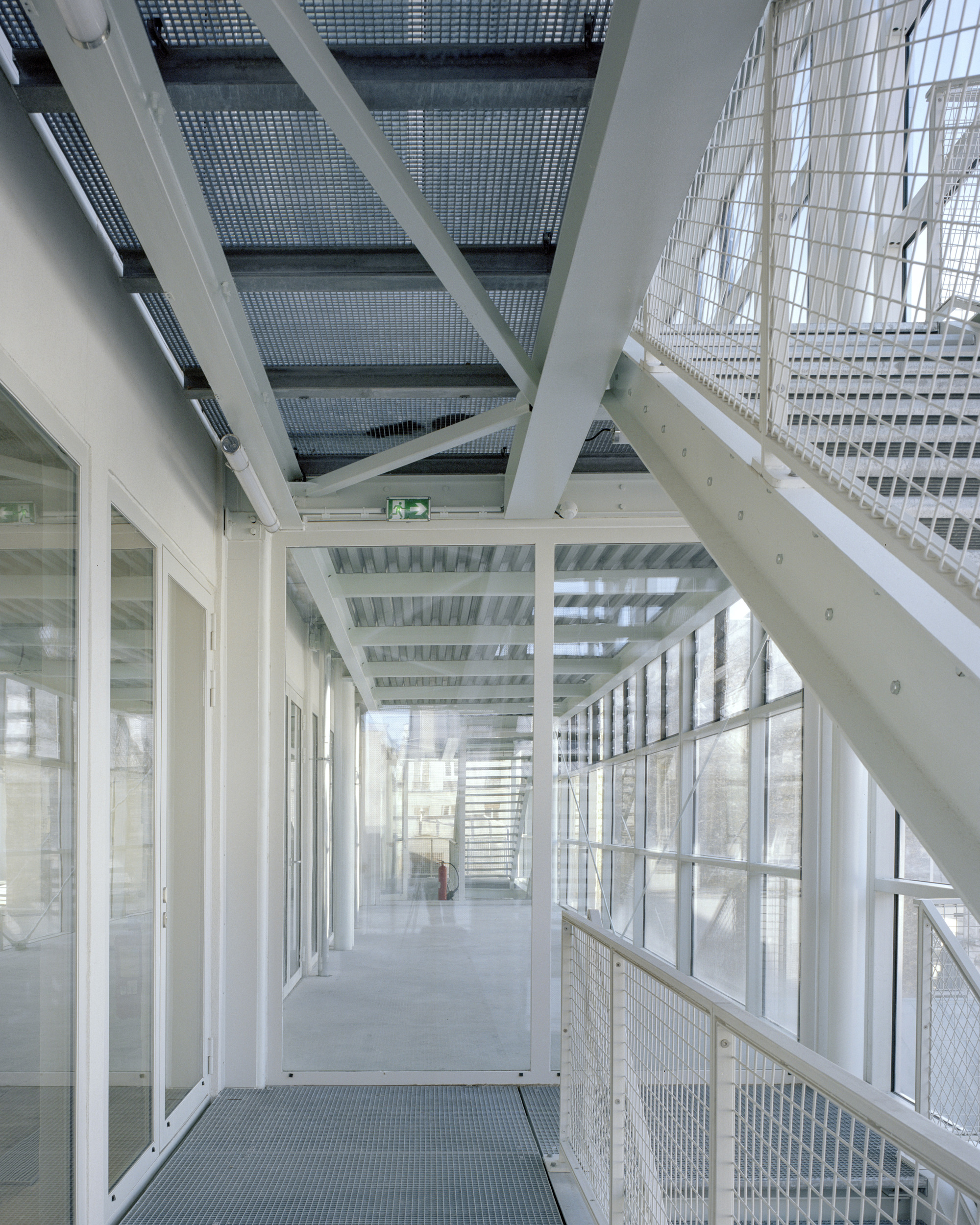 Interior steel walkway with grating floors, white diagonal braces and a mesh-railed stair rising beside glazed walls in soft daylight