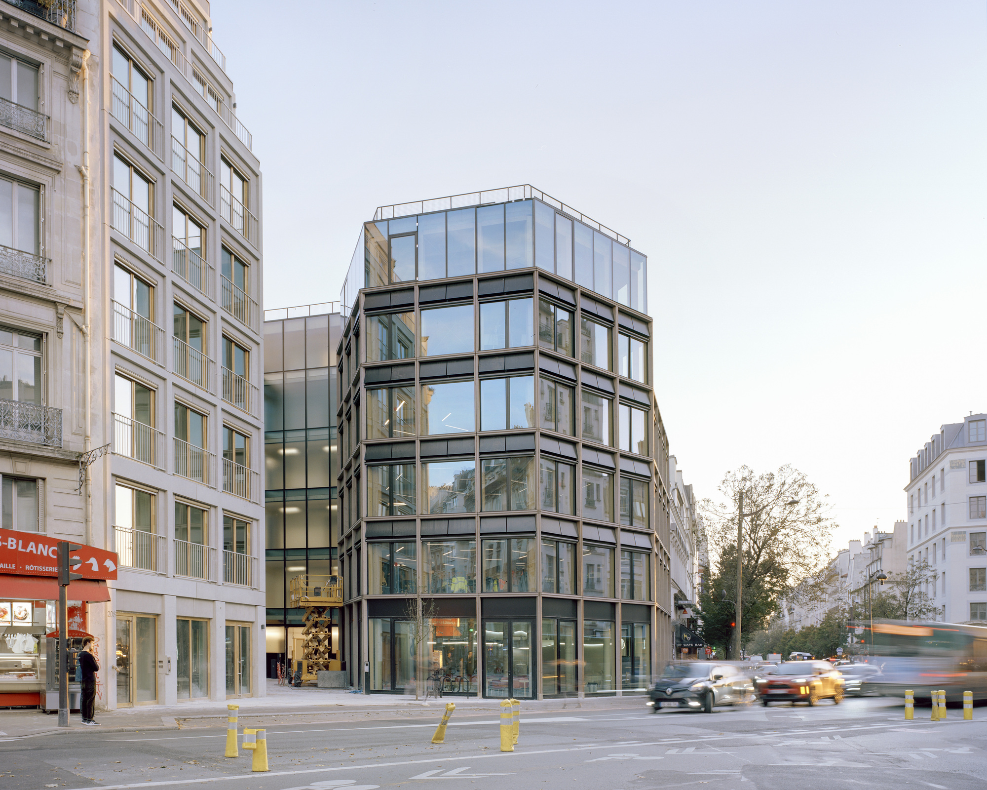 Street corner view of a dark framed glass building beside a stone facade, with cars streaking past in blurred motion at dusk