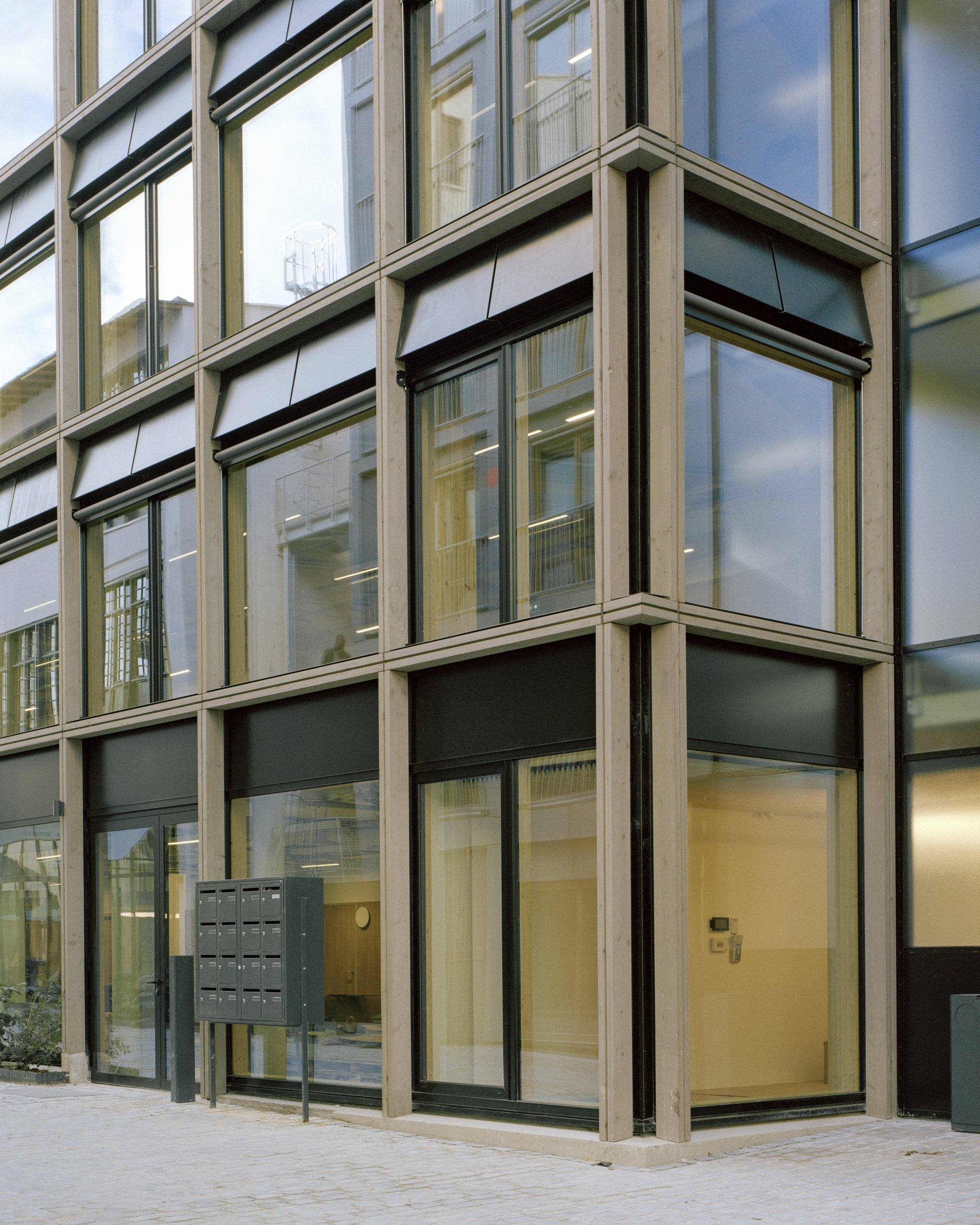 Corner of a timber-framed glass facade with dark awnings and a mailbox cluster beside the entrance at dusk