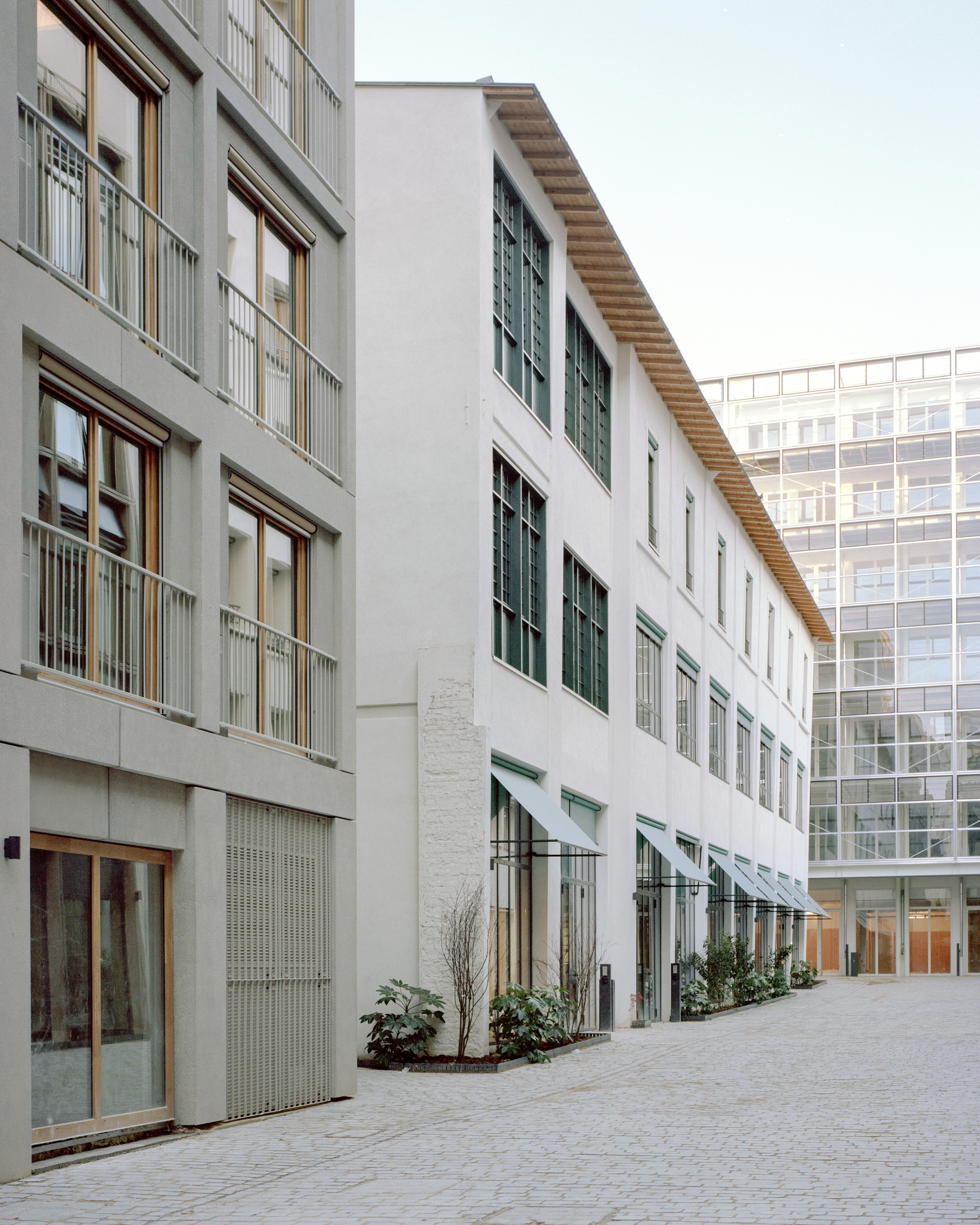 Cobbled courtyard lined with white rendered facades, green window awnings and a slatted timber eave beside a glass tower in soft light