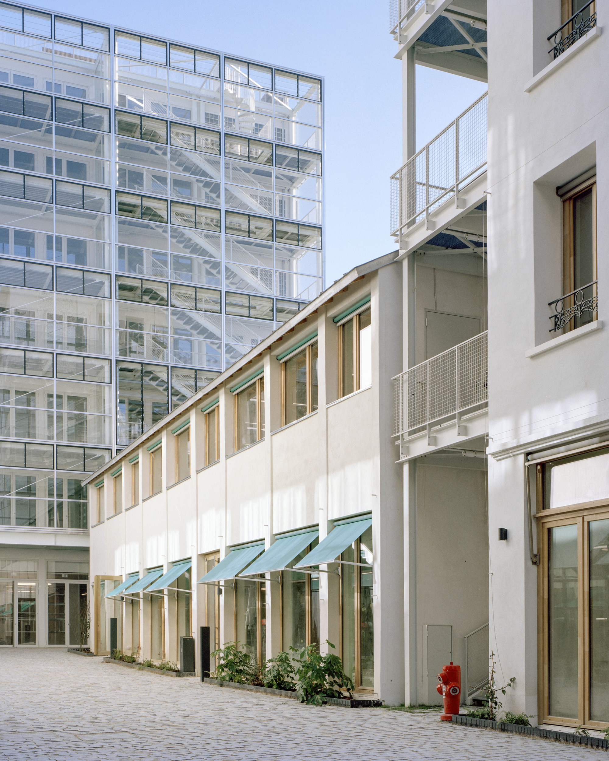 Low white building with teal awnings and planted beds beside a glass tower revealing stairs, a red hydrant in the foreground
