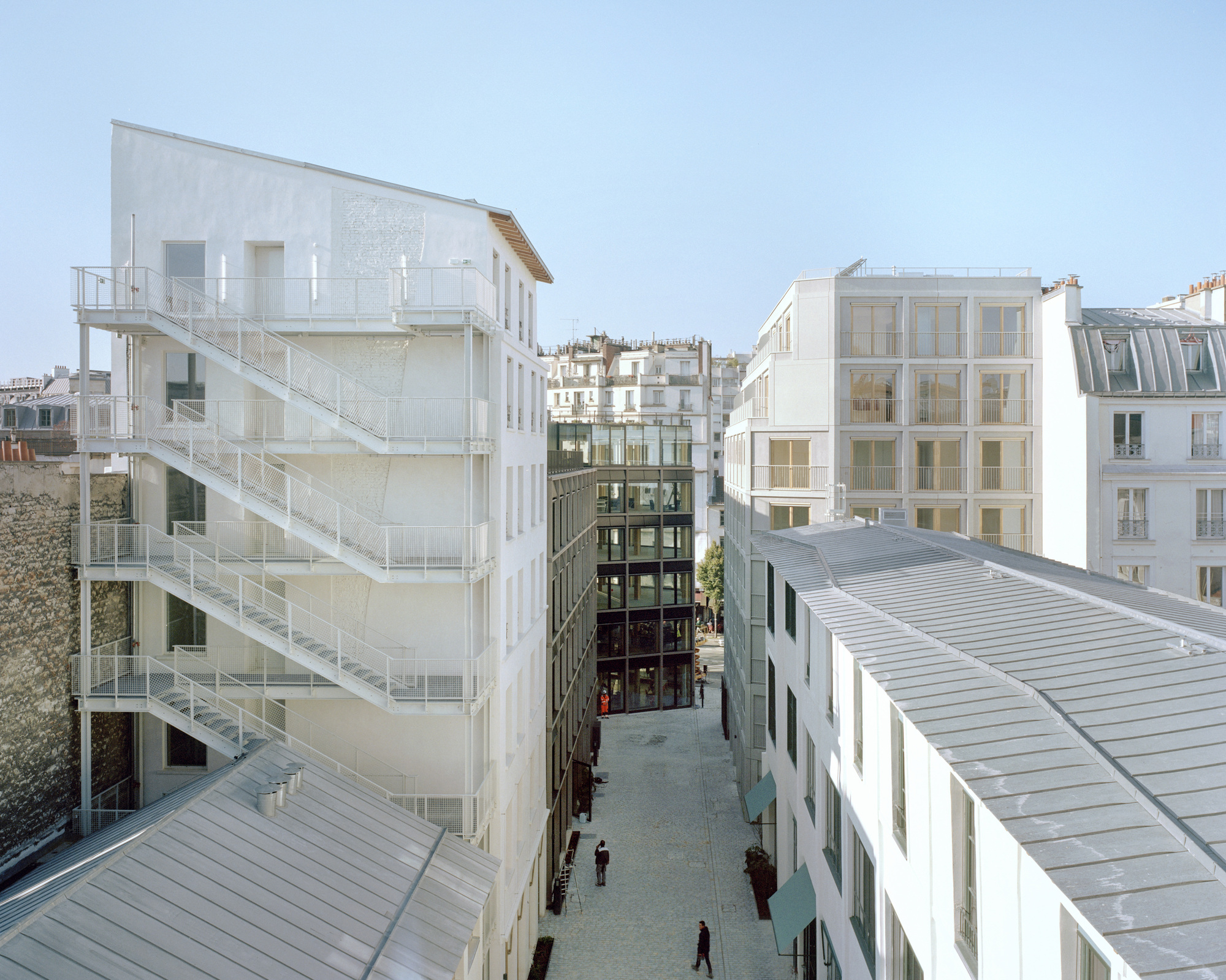 White building with zigzag external steel stairs above a narrow pedestrian lane, with two people walking between zinc roofs
