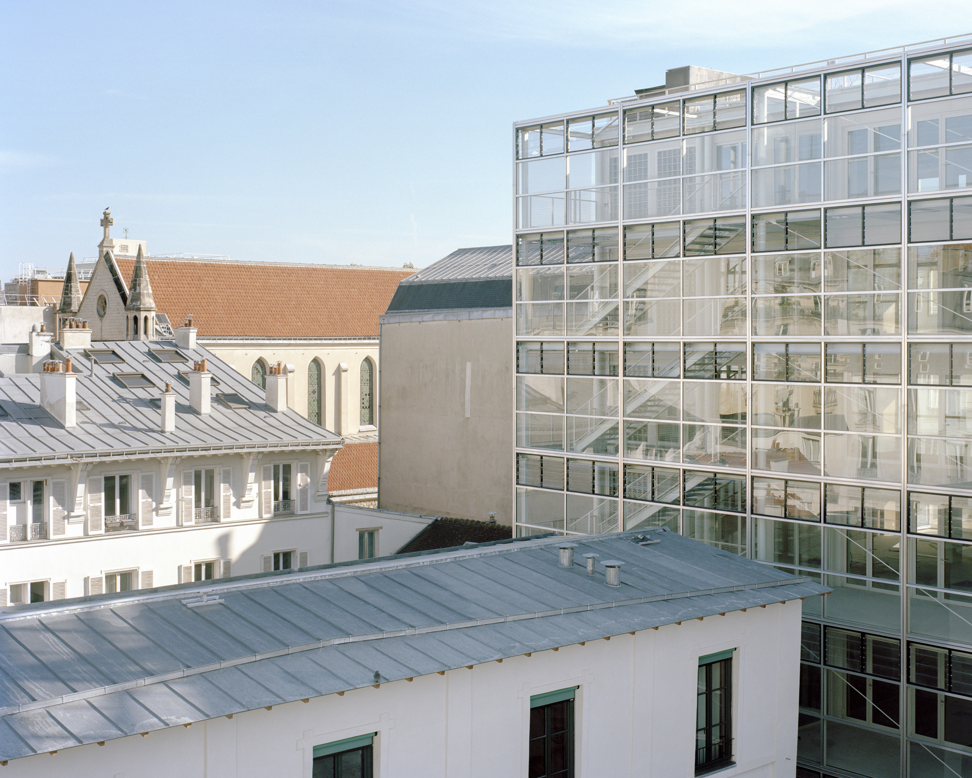 Glass-walled stair tower rising beside zinc roofs, chimneys and a pitched tile roof church under a pale blue sky