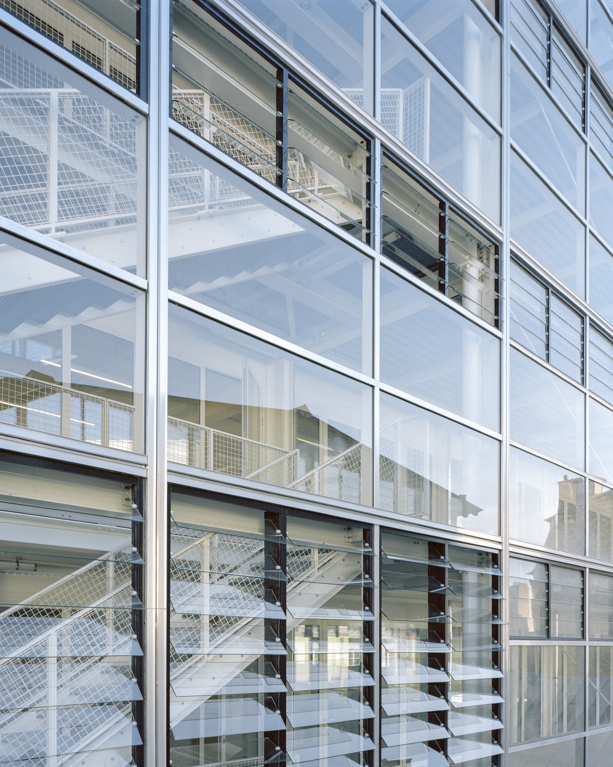Close-up of the glass curtain wall with operable louvre windows, revealing mesh-railed steel stairs behind the glazing in soft daylight