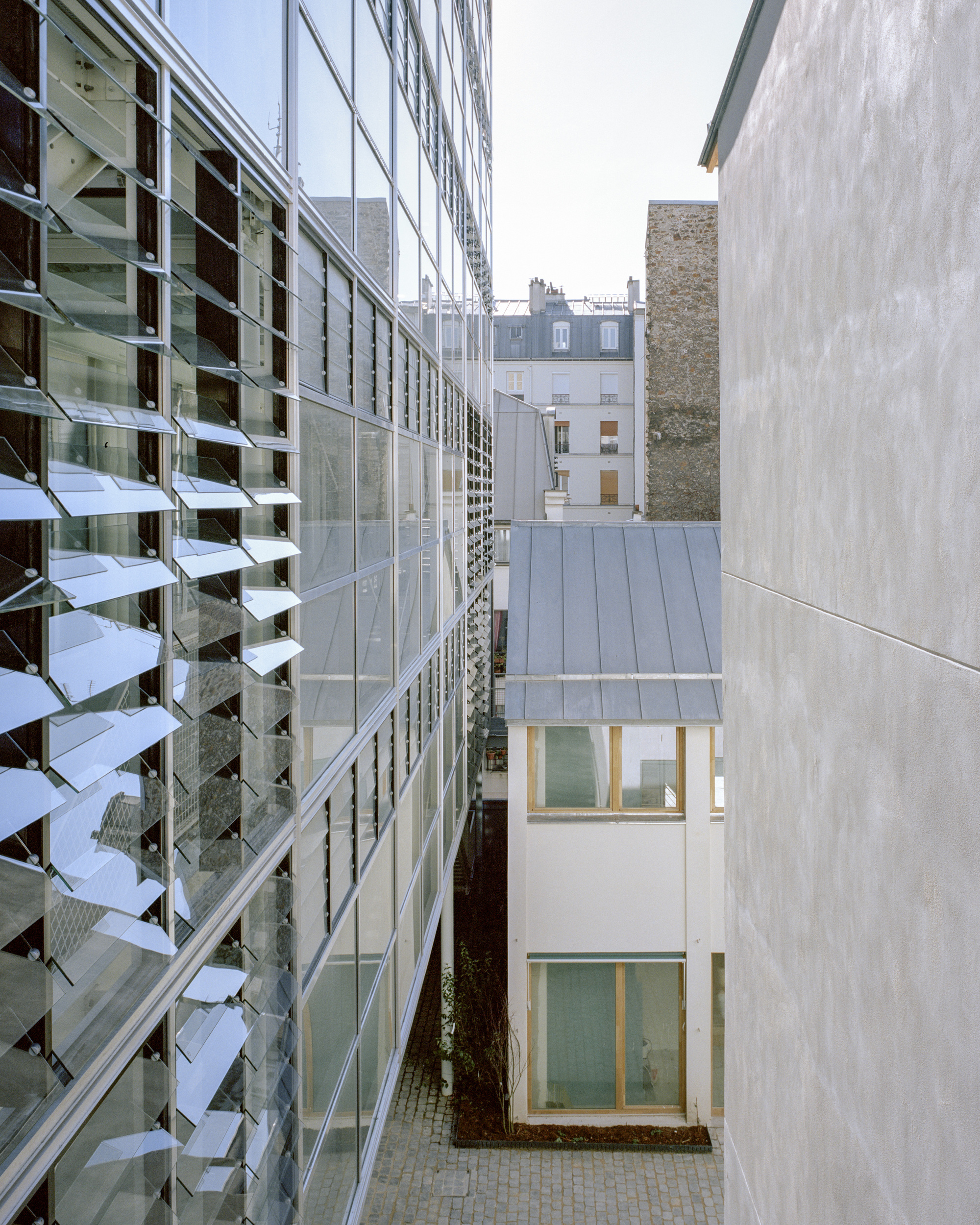 Narrow gap between the louvred glass facade and a pale concrete wall, with a zinc-roofed block and cobbled courtyard below