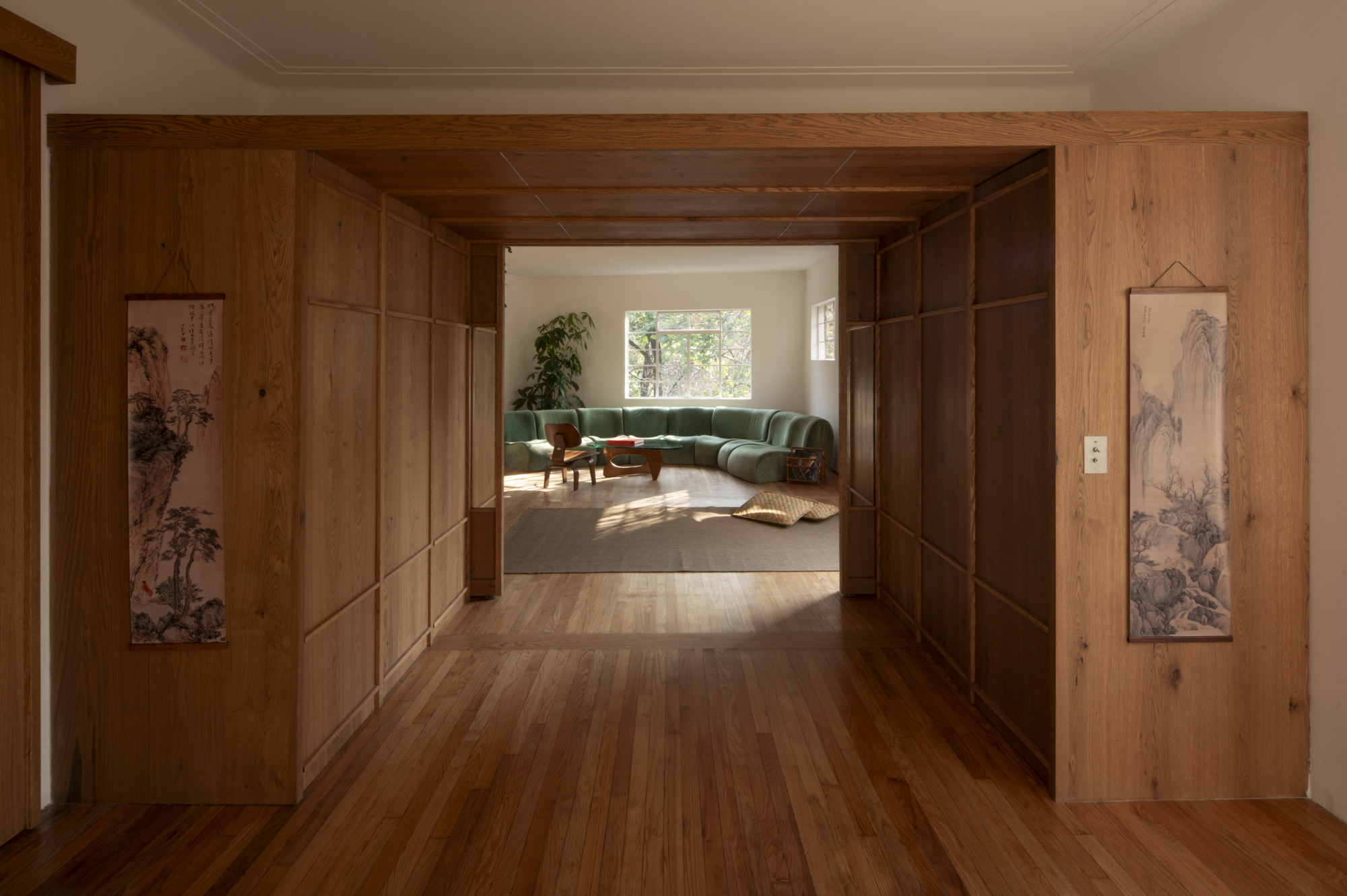 Oak-paneled threshold framing a curved green sofa and sunlit rug, with two hanging ink scroll paintings flanking the opening