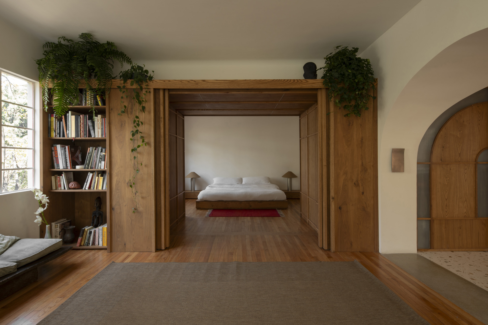 Bedroom seen through a timber portal with sliding panels, bookshelf and trailing ferns beside a window with leafy light