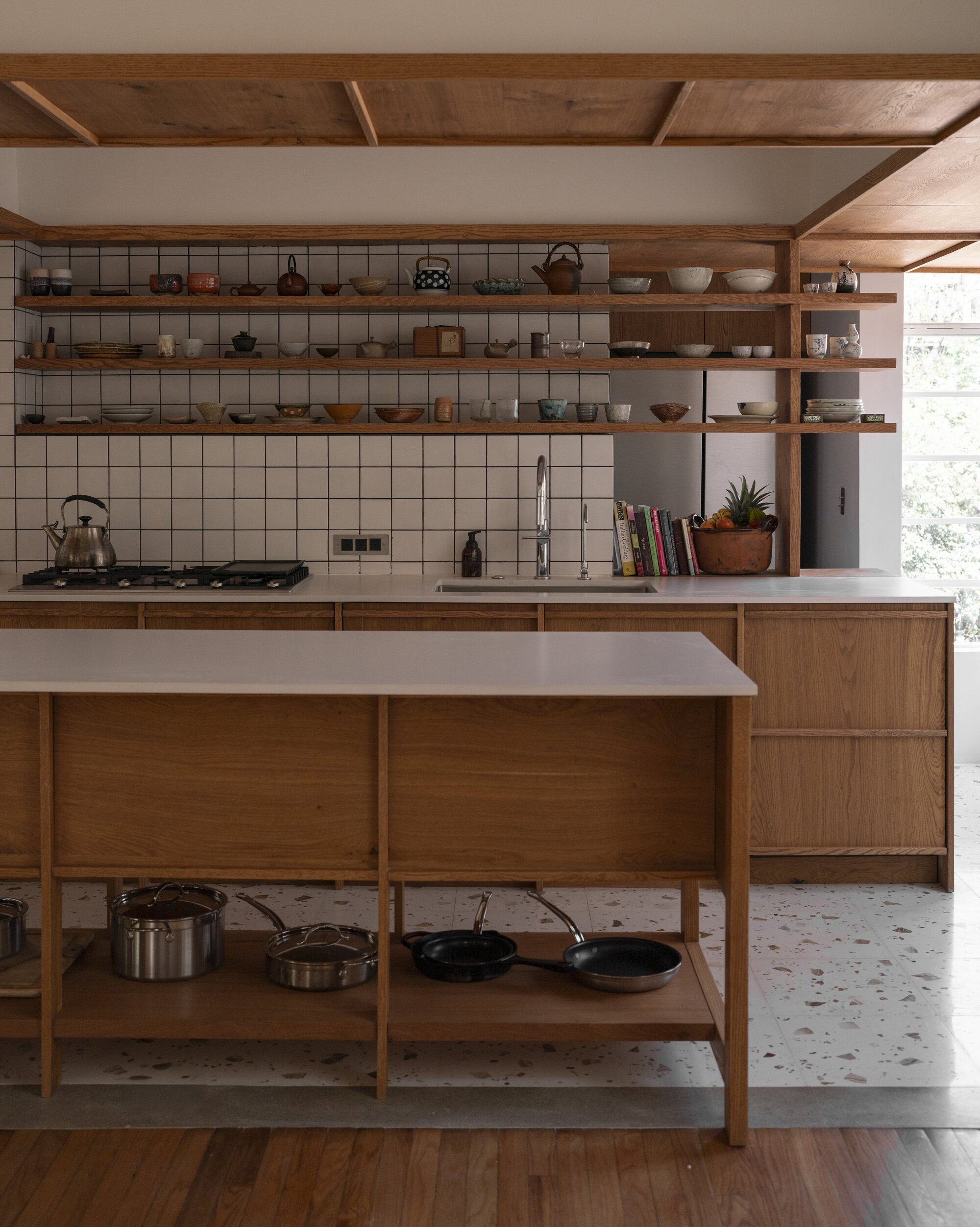 Oak kitchen island and open shelving of ceramics against white grid tile, with terrazzo floor and daylight from a side window