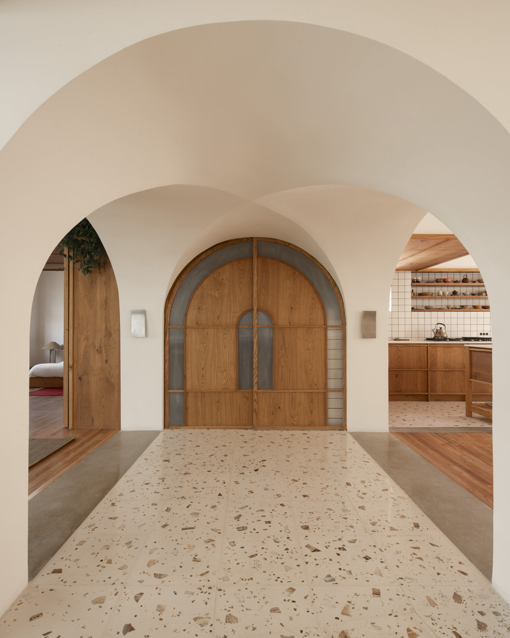 Plastered arches leading to an arched wooden door with glass inserts, over a speckled terrazzo floor with the kitchen glimpsed beyond