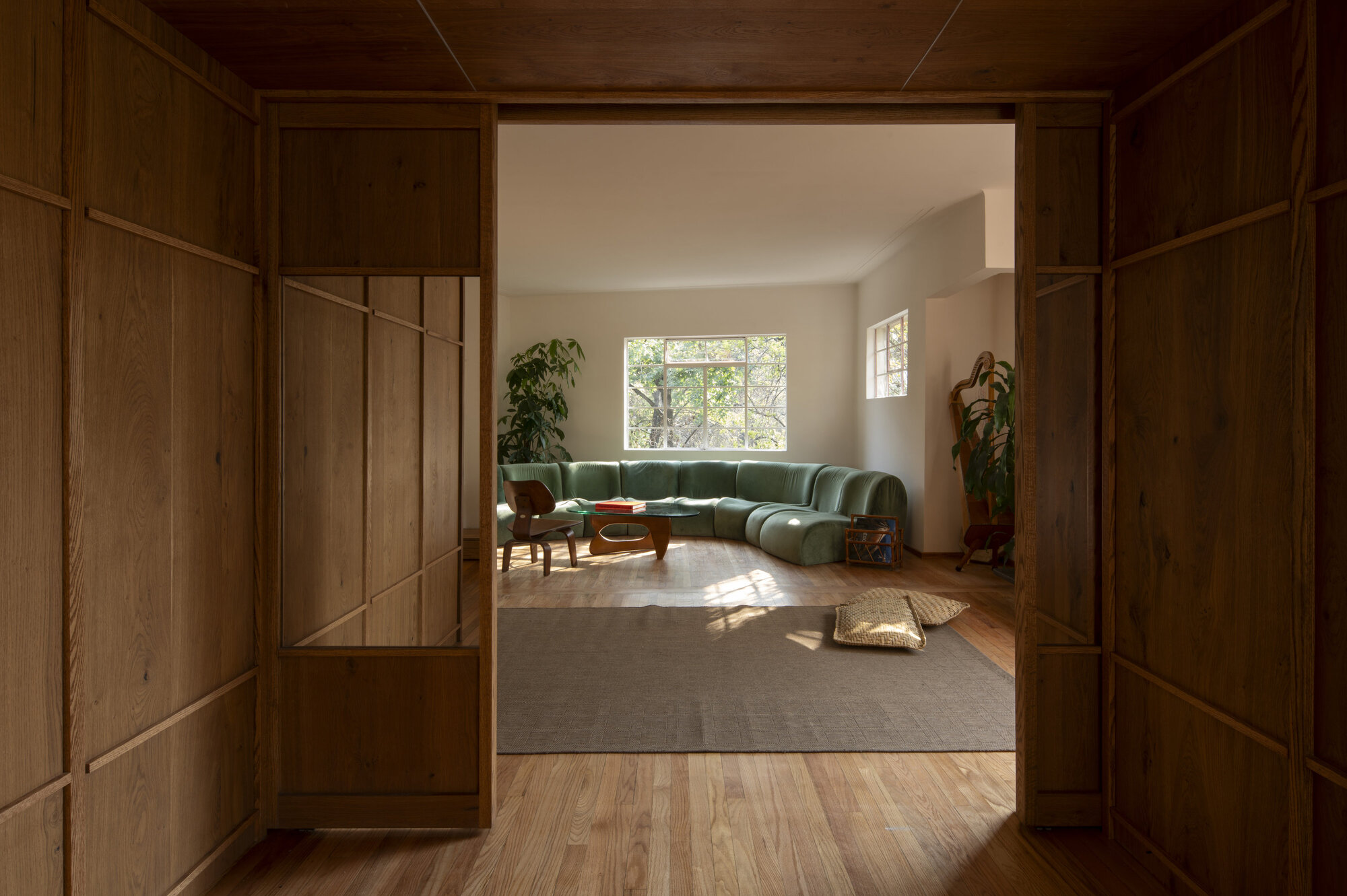 Dark oak panel doorway opening onto a green curved sofa, woven rug and sunlit window with trees outside
