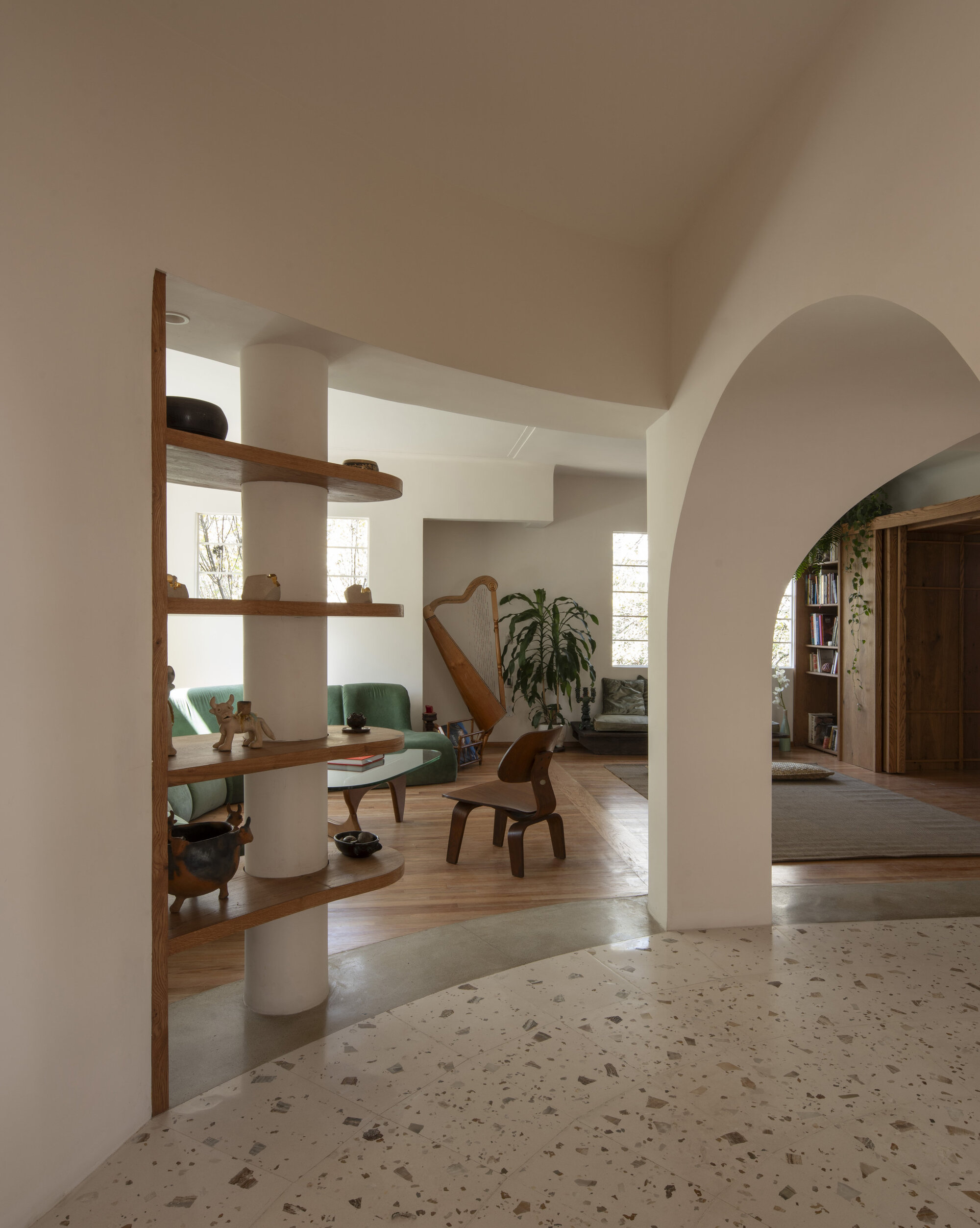 Curved timber shelves wrapped around a white column beside a plaster arch, with terrazzo floor and green sofa in soft daylight