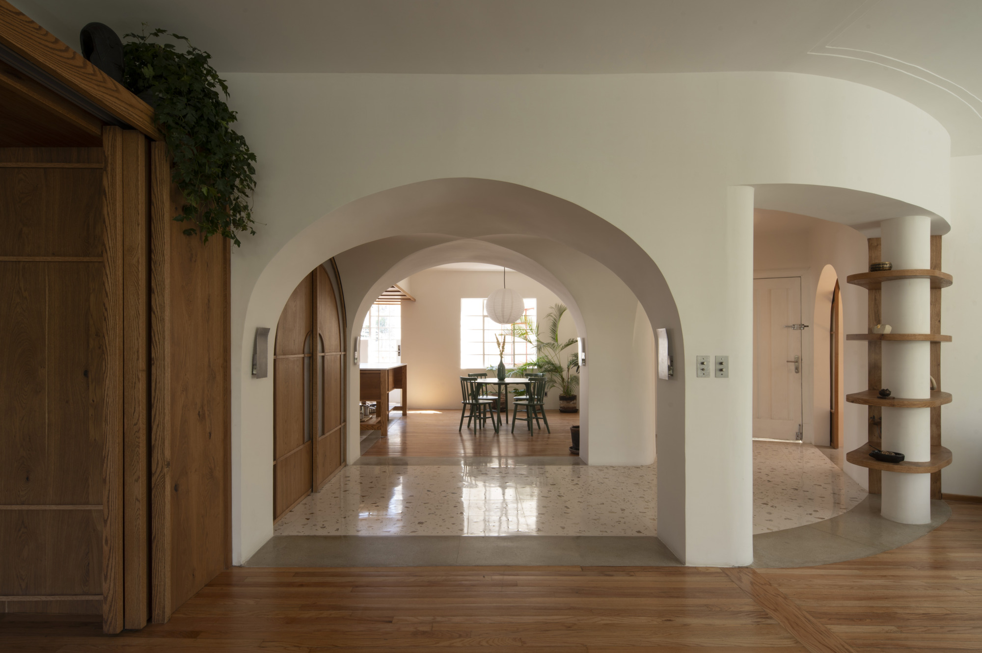 Enfilade of plaster arches leading from a wood floor to a terrazzo dining room, with a planted timber door at left
