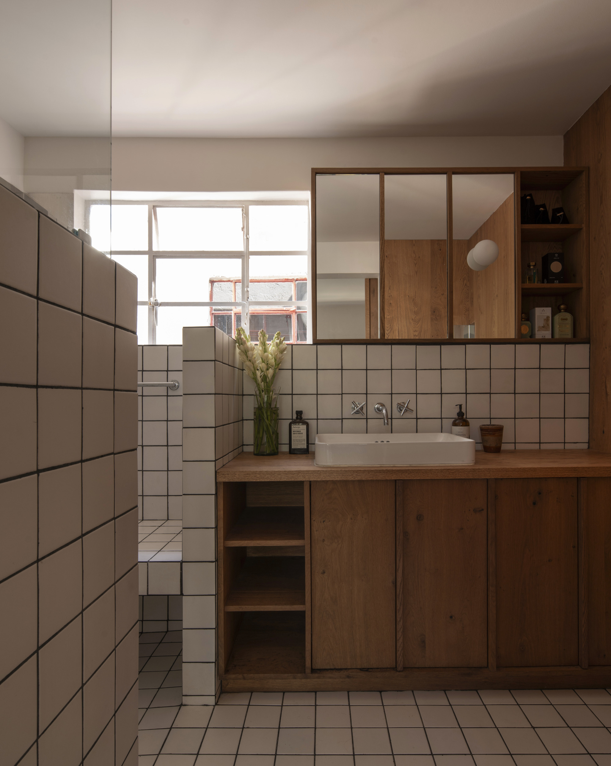Timber vanity with a white basin beneath a mirrored cabinet, with a steel-framed window casting daylight over grid tile walls