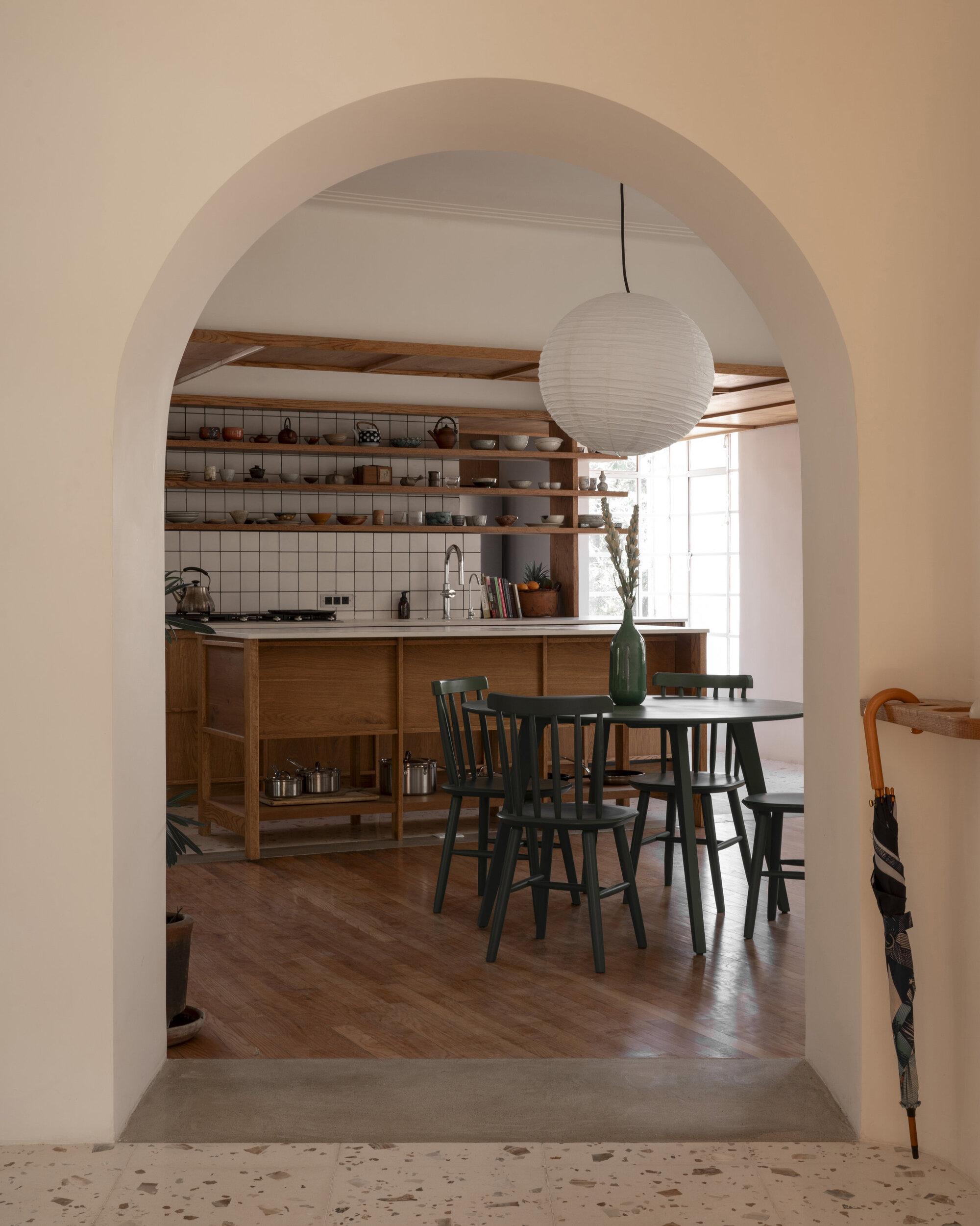 Arched opening framing a round dining table with green chairs and a paper pendant lamp, kitchen island beyond, umbrella at right