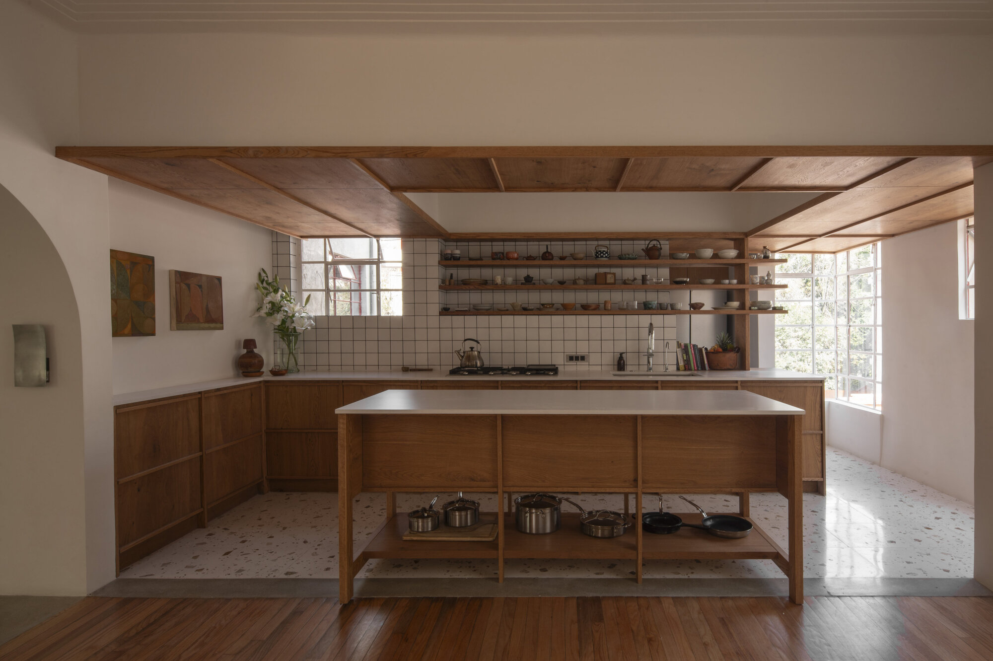 Kitchen with a timber island, open shelves of ceramics and a wood-panelled ceiling canopy, bright steel windows at right