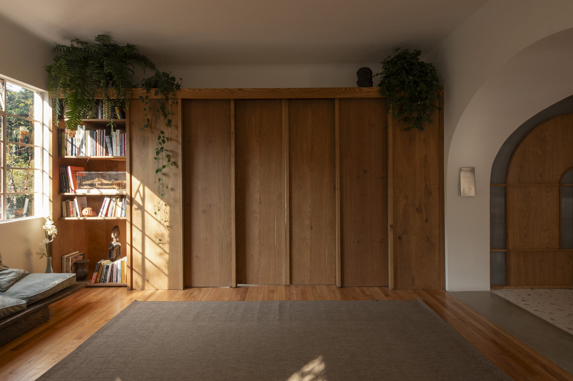 Wall of sliding oak panels beside a built-in bookshelf, with ferns on top and low sun across the wood floor