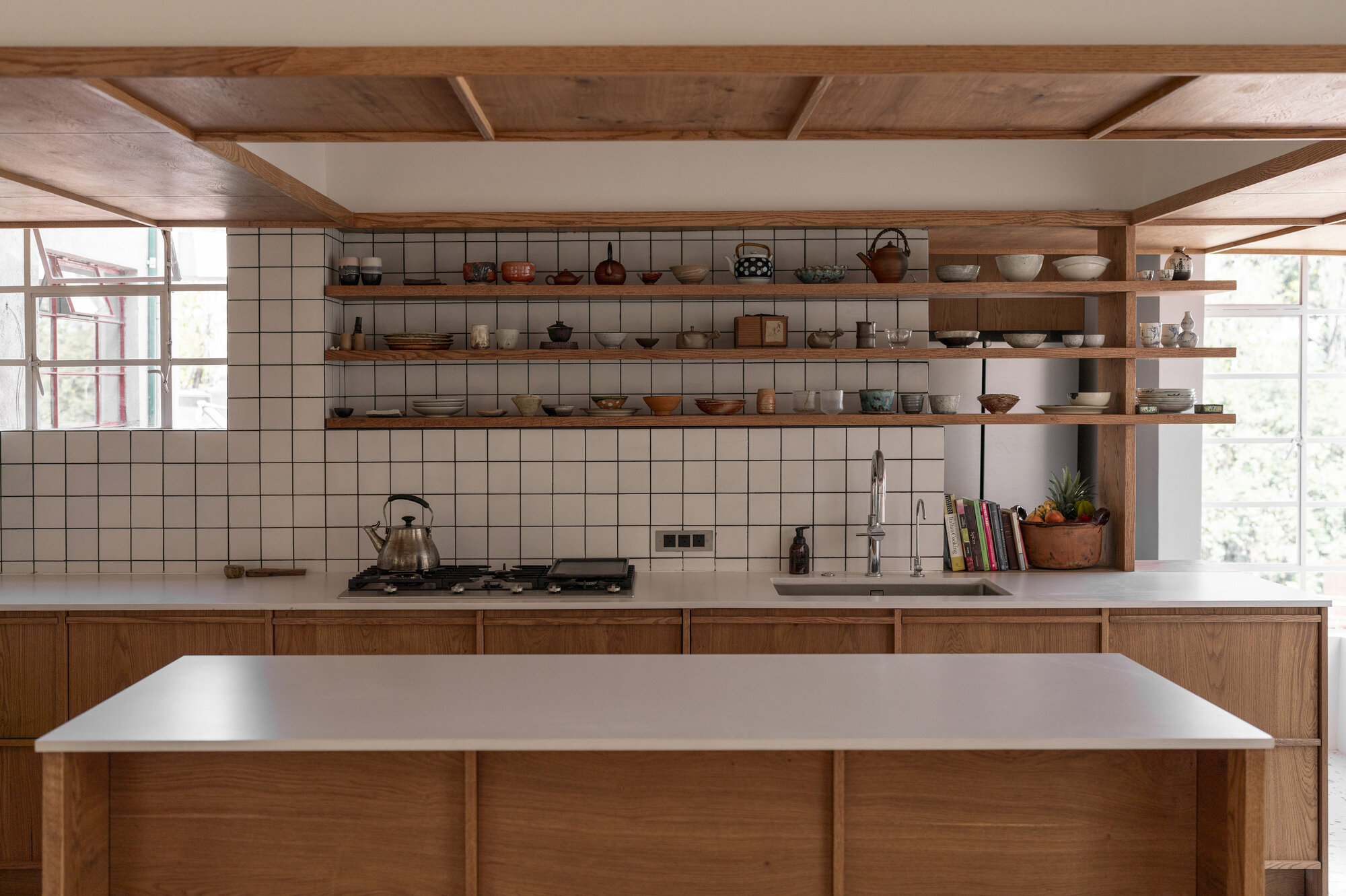 Kitchen with open timber shelves of ceramics over white grid tile, an oak island in the foreground and daylight from corner windows