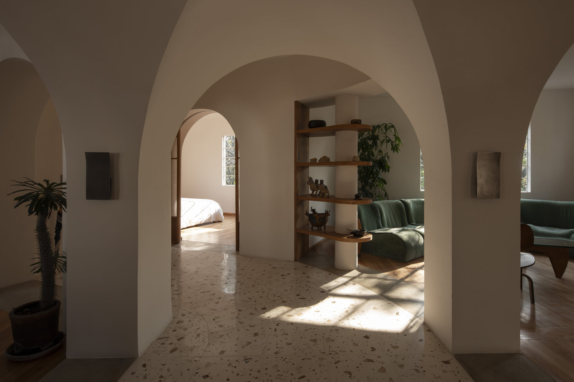 Rounded white archways opening onto a terrazzo floor, with sunlight patches and a green sofa visible in the next room