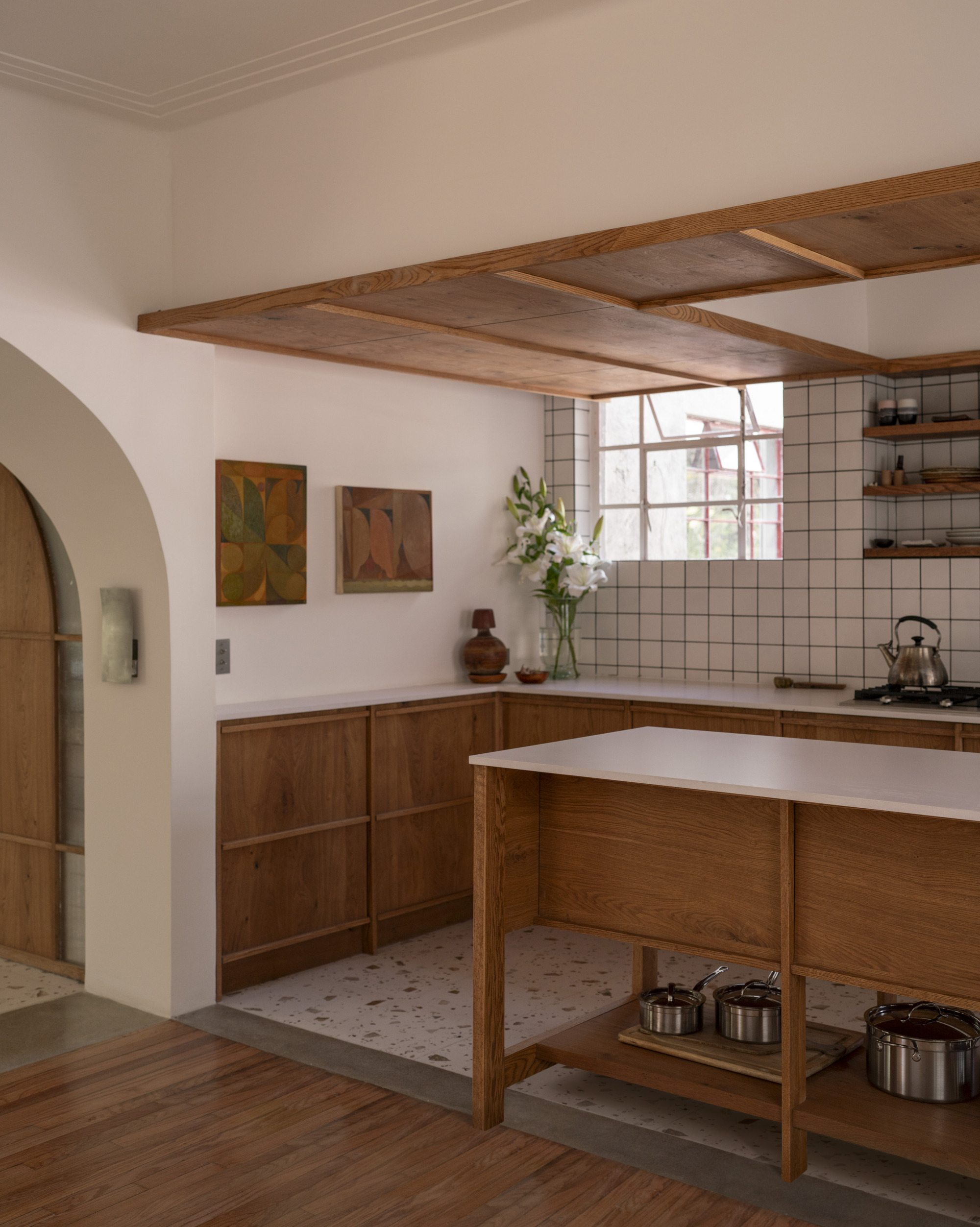 Kitchen corner with oak cabinets, a timber ceiling frame over the counter and a vase of lilies by the window
