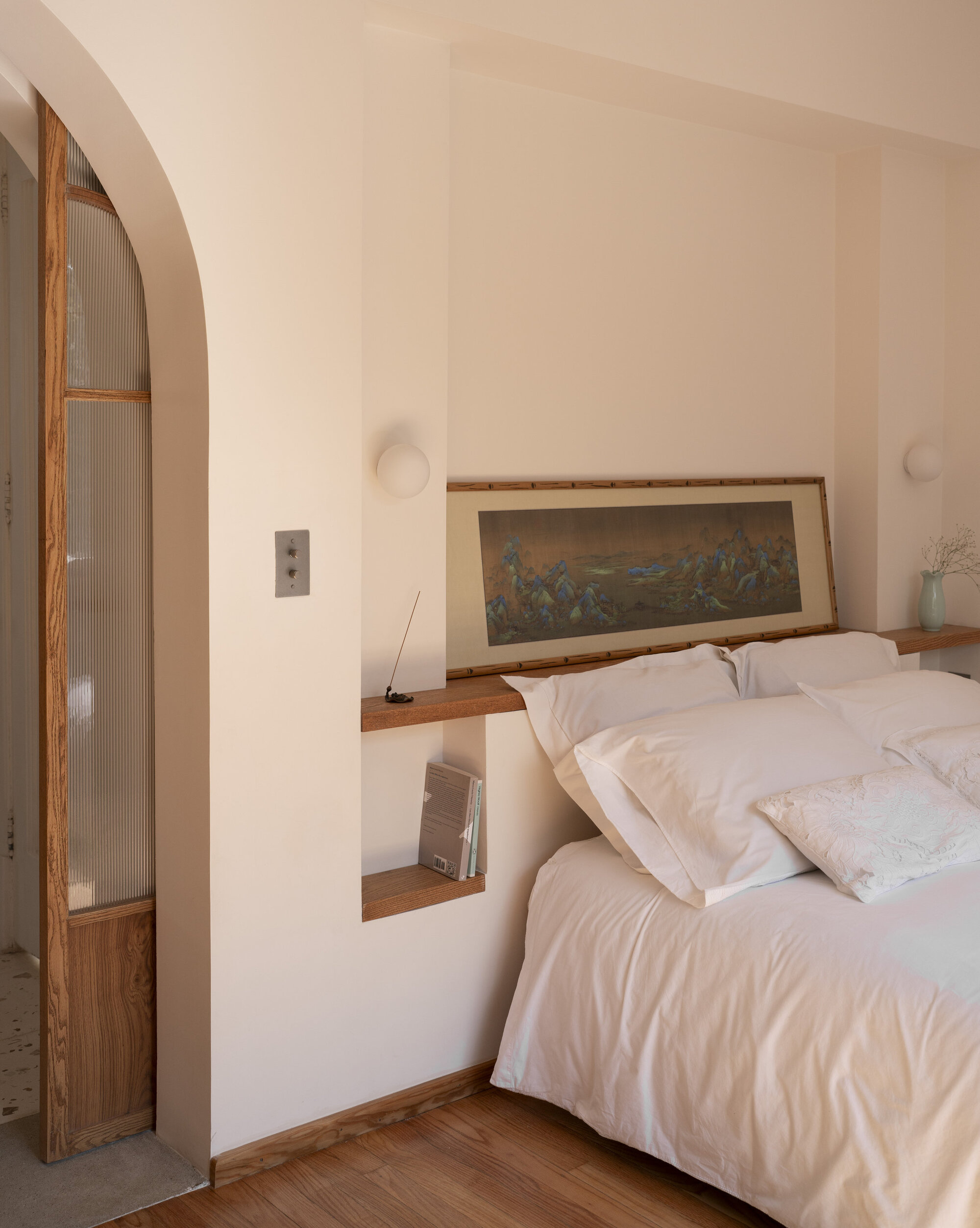 Bedroom alcove with a built-in timber shelf behind the bed, a framed landscape scroll and a fluted glass door in an arch