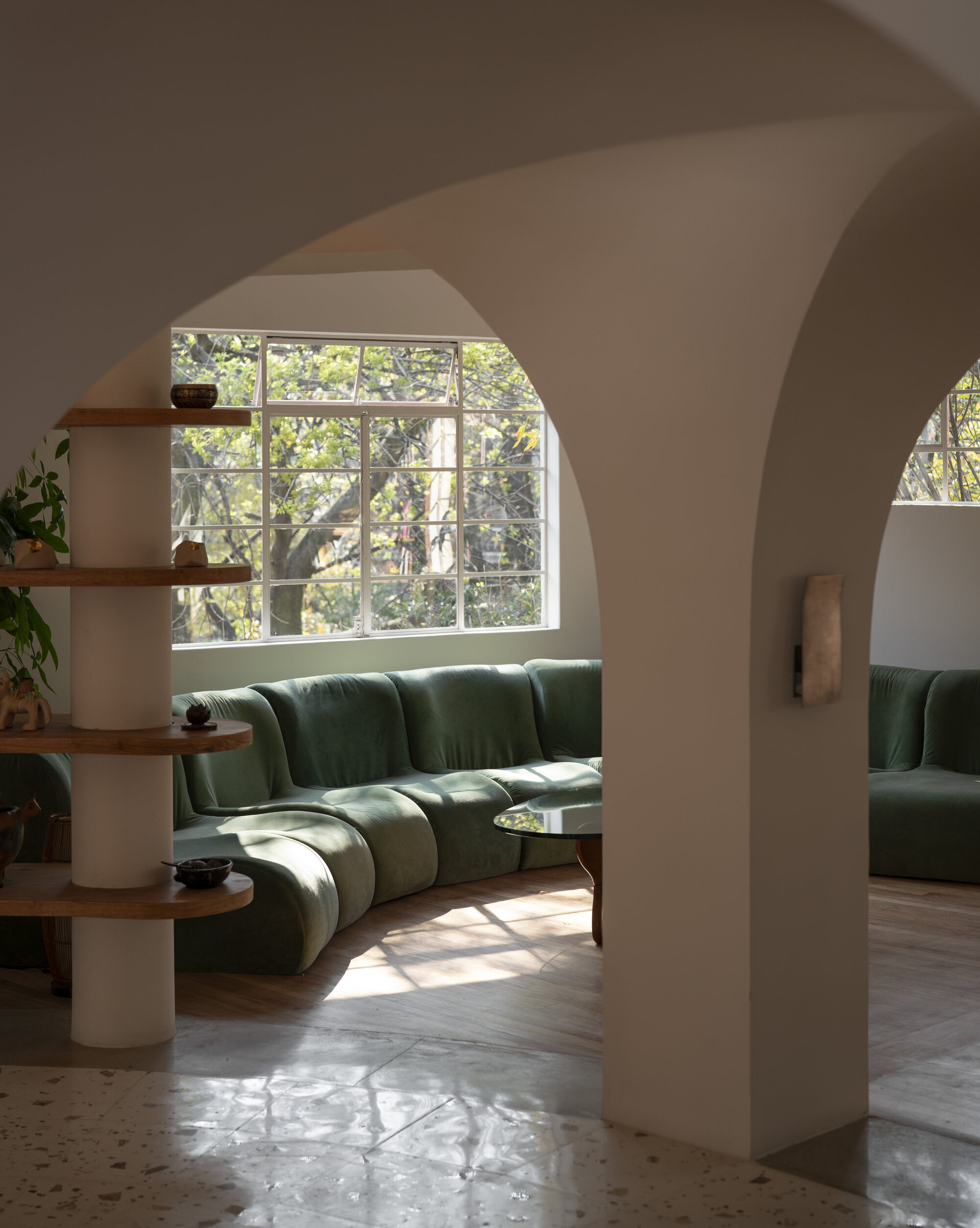 Curved green velvet sofa under a tall gridded window, framed by plaster arches and a round shelving column with trees outside