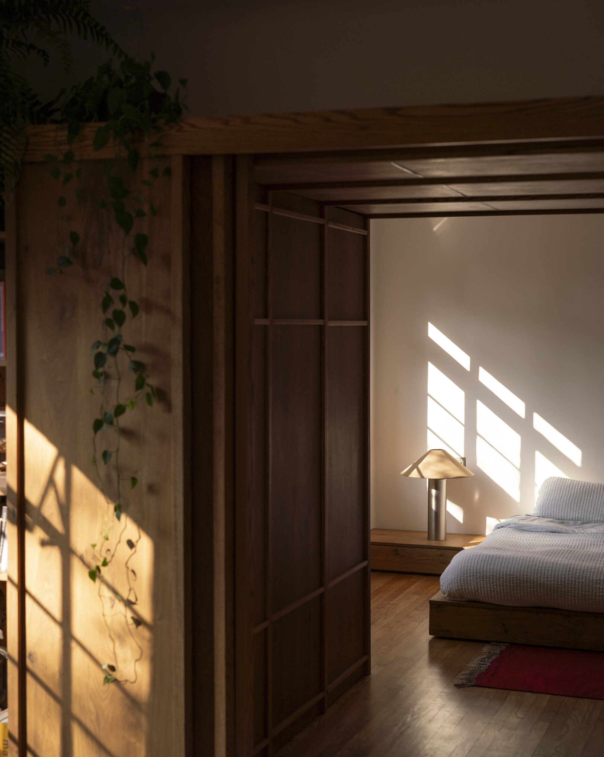 Timber-framed doorway opening onto a low bed and table lamp, with window shadows cast across the wall in low evening sun