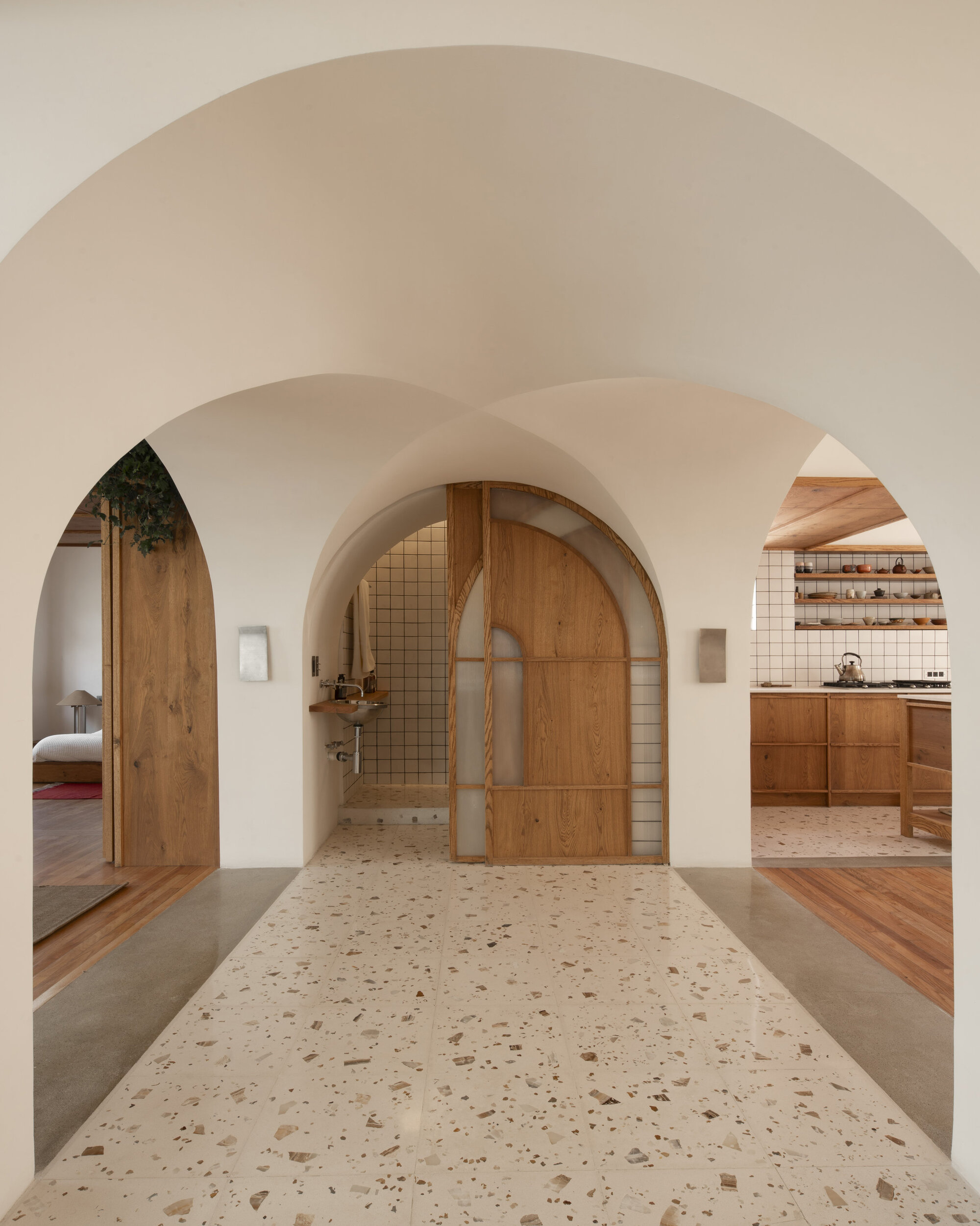 Succession of white plaster arches over a terrazzo floor, with an arched timber and glass door and kitchen beyond, in soft daylight