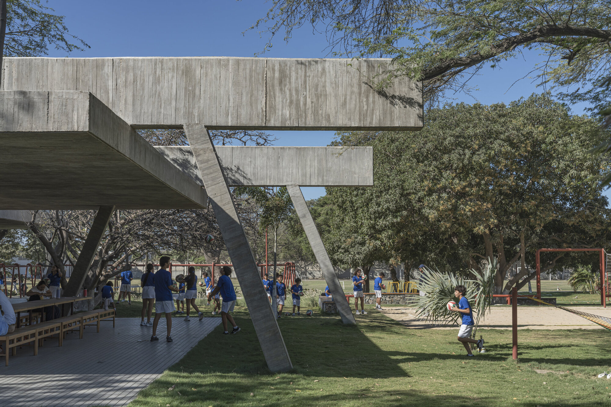 Slanted concrete columns and cantilevered beams over a timber deck, with children in blue shirts playing on the lawn in afternoon sun