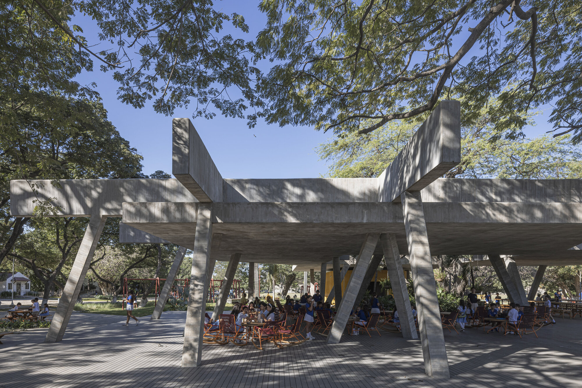 Board-formed concrete canopy on angled columns sheltering people at tables, with tree branches overhead and dappled shade on the deck