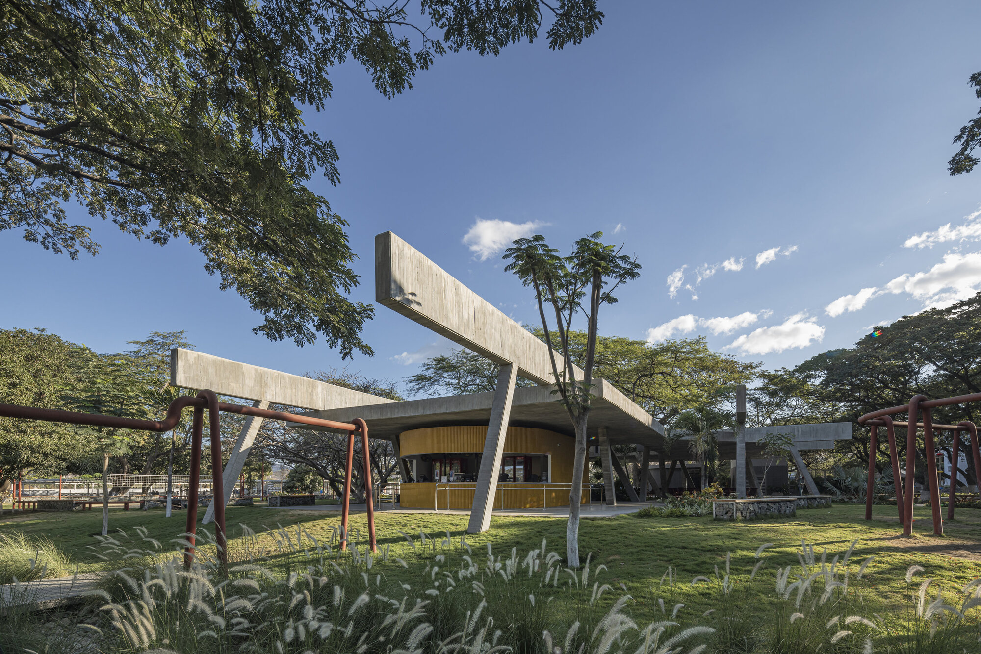 Concrete canopy with long projecting beams above a yellow curved kiosk, seen past tall grasses and red pipe play structures under blue sky
