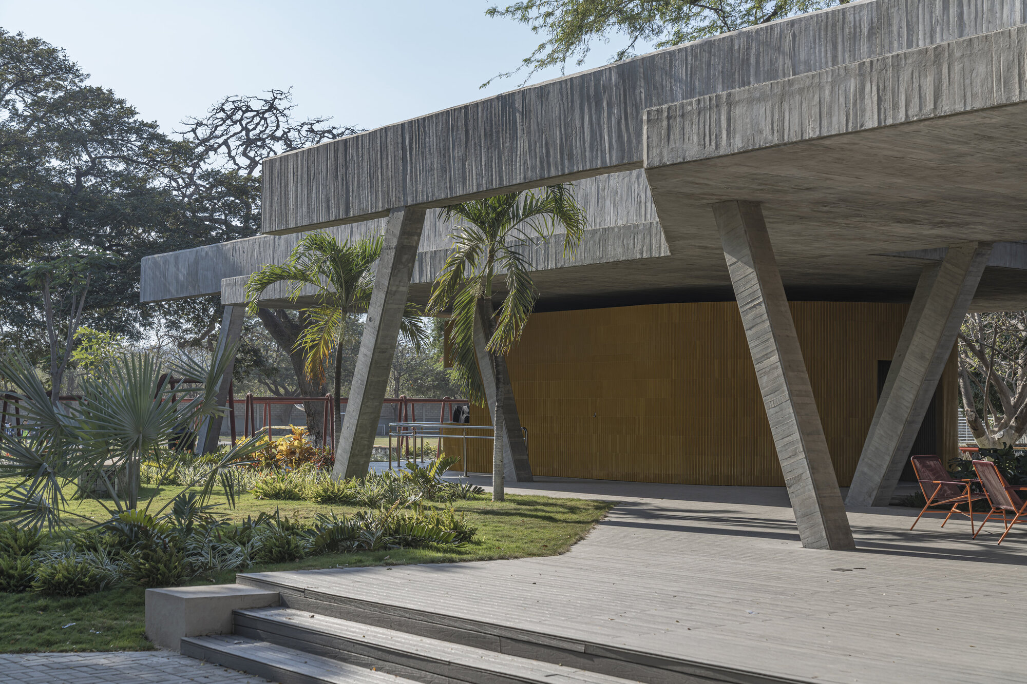 Angled concrete columns and board-marked beams above a curved yellow slatted wall, with palms and orange chairs on a timber deck