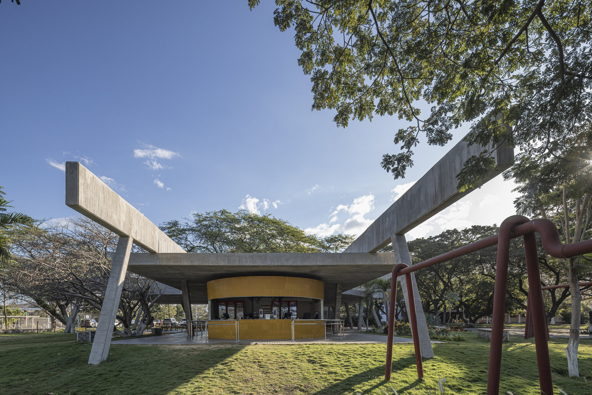 Low-angle view of the concrete canopy with upswept beams over a yellow curved kiosk, framed by a red pipe structure and tree leaves