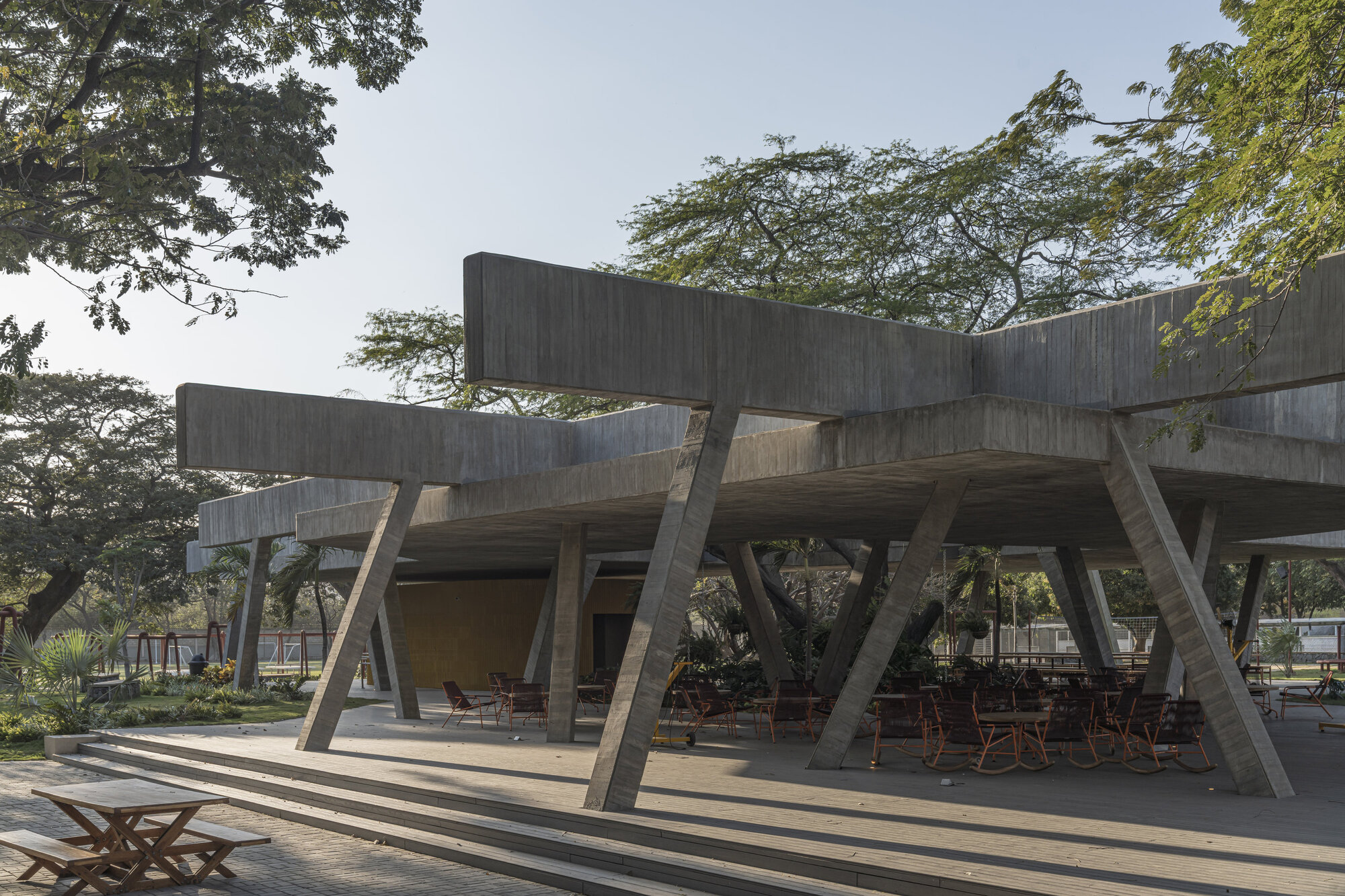 Raised concrete canopy on slanted columns above a timber deck with red chairs, framed by large shade trees in hazy sun