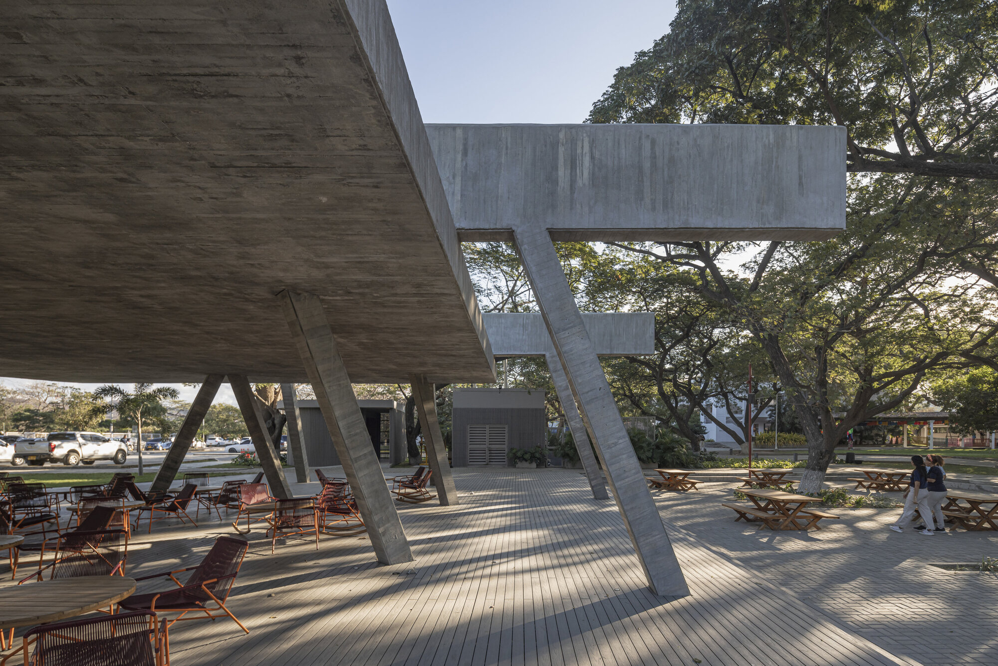 Angled concrete columns holding a cantilevered slab over a wooden deck, with two people walking past picnic tables in low light