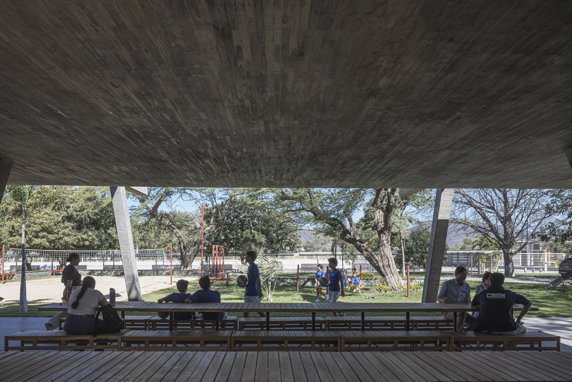 View from under the board-marked concrete soffit across long timber tables toward children playing on a sunlit lawn