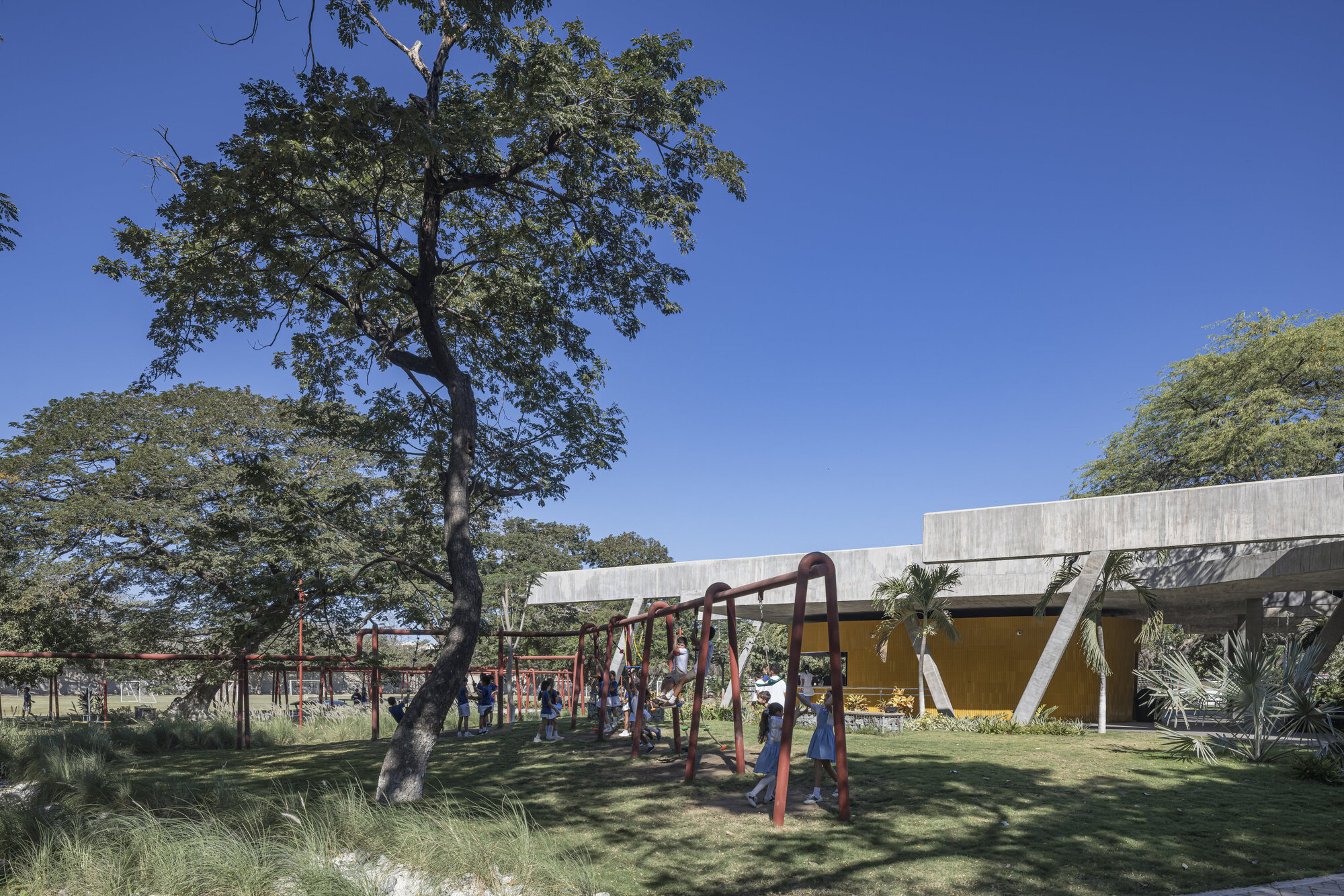 Children on a red steel swing frame in a grassy yard, with the concrete roof edge and ochre wall behind under deep blue sky