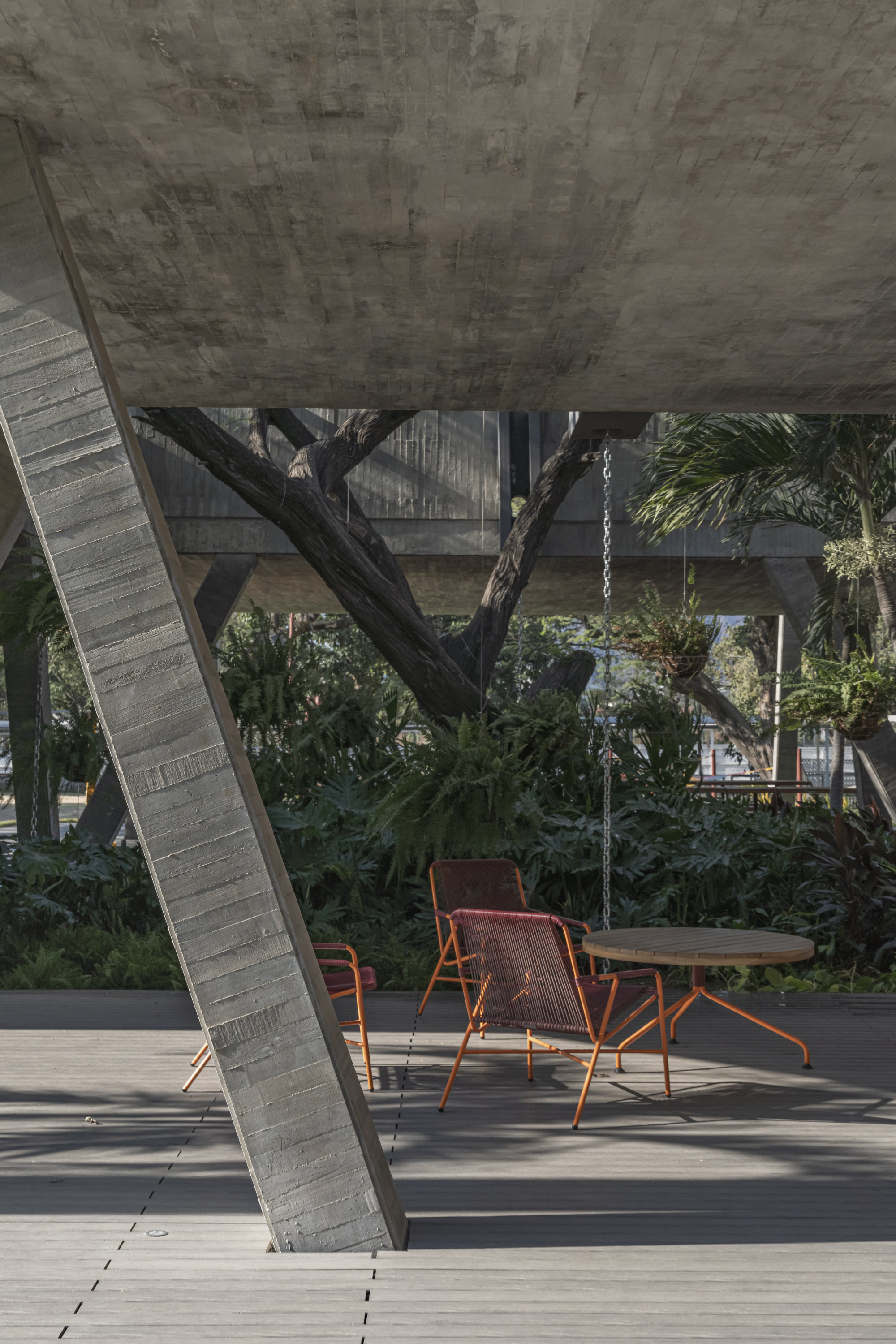 Board-marked concrete column leaning over a timber deck with orange chairs and a round table, ferns and a tree trunk beyond