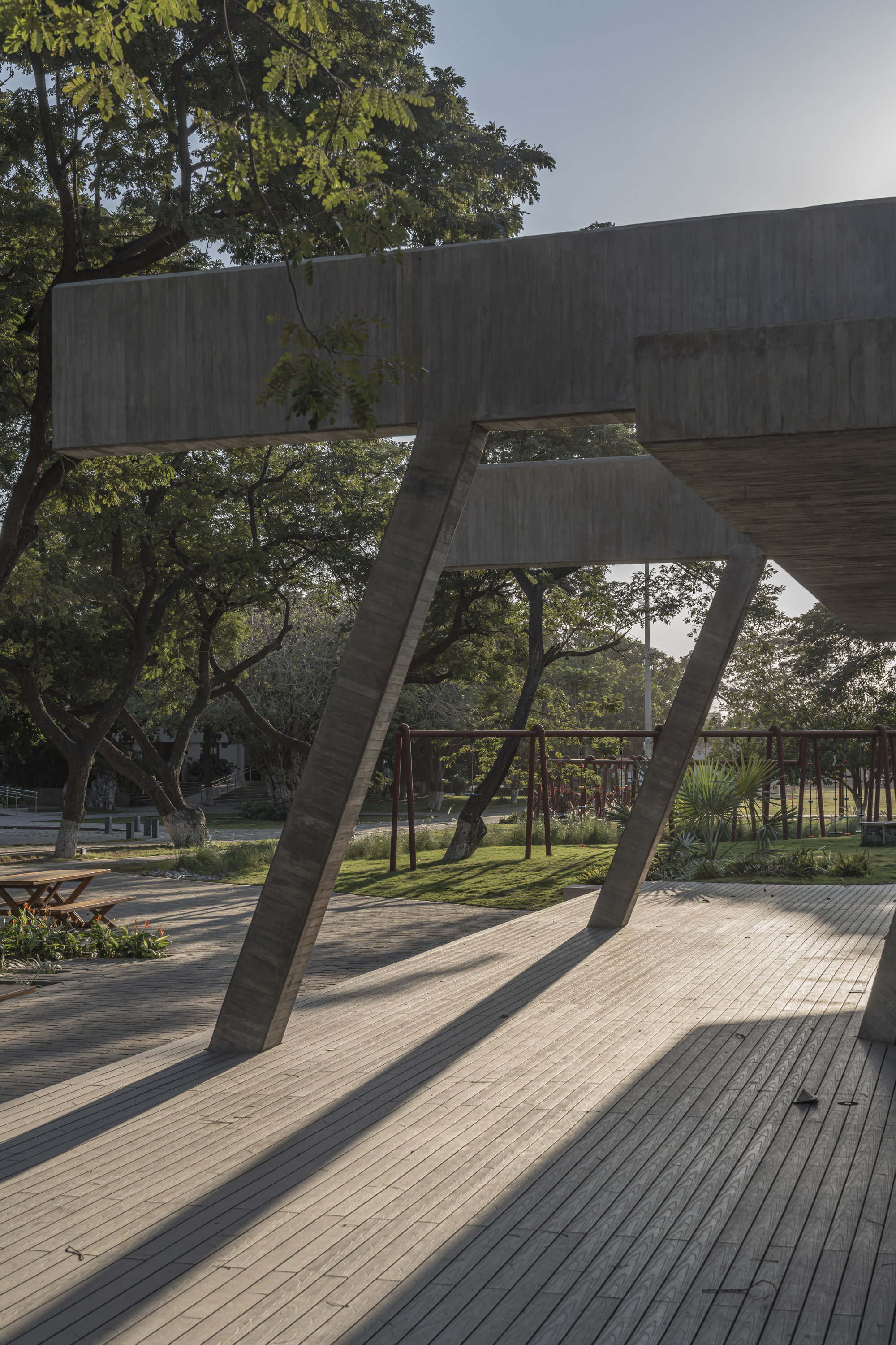 Slanted concrete columns holding a deep beam above a timber deck, with long late-afternoon shadows across the boards