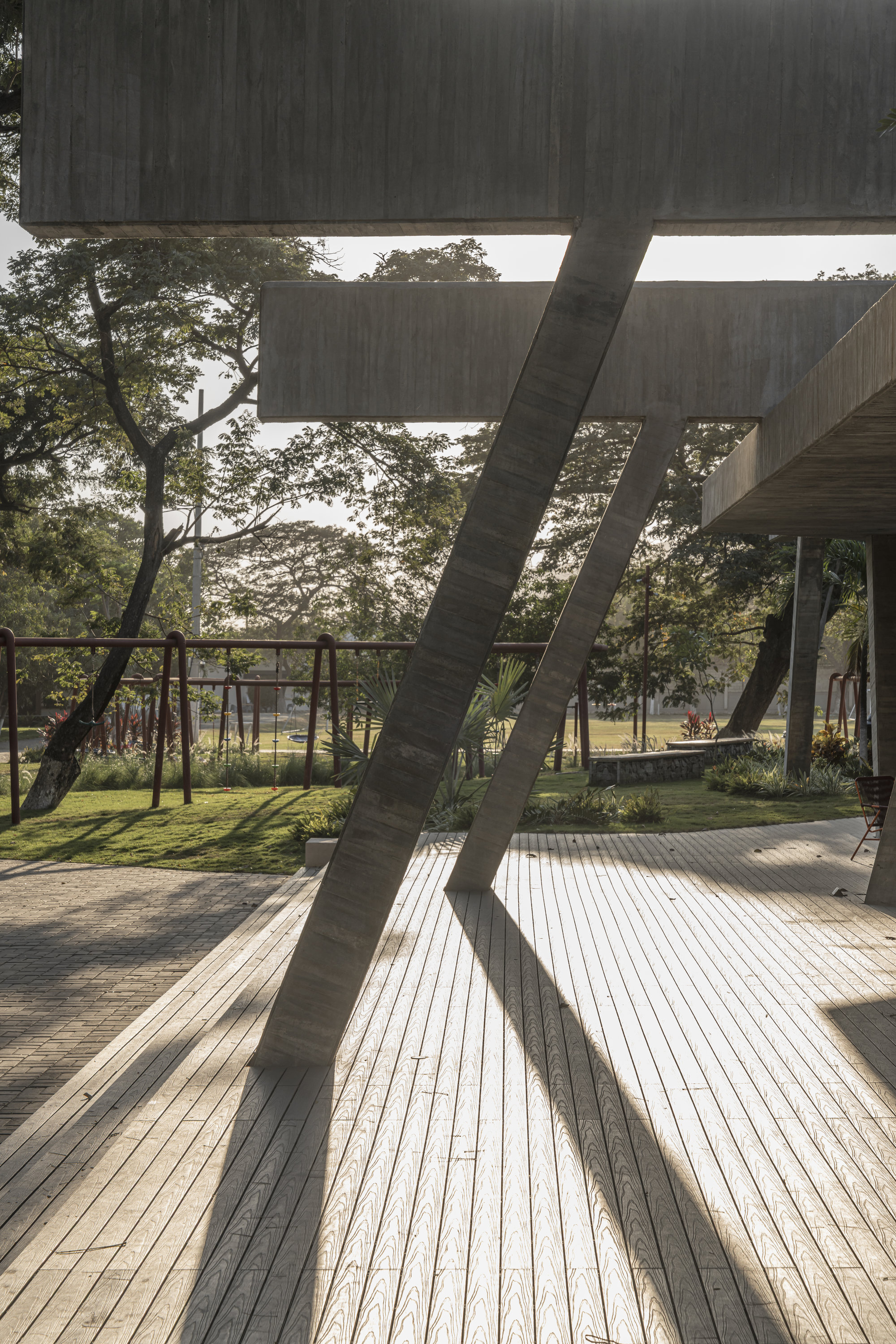 Pair of diagonal concrete struts under stacked beams, their shadows stretching across a timber deck toward a grassy lawn