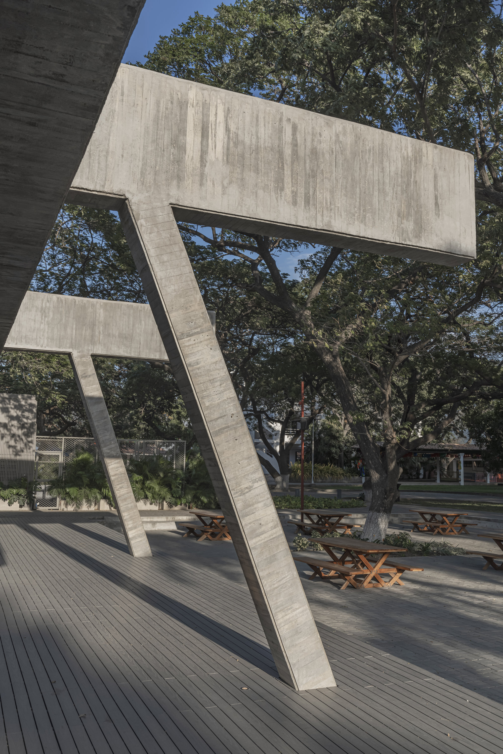 Tilted concrete column and cantilevered beam above a deck with wooden picnic tables, set among large shade trees