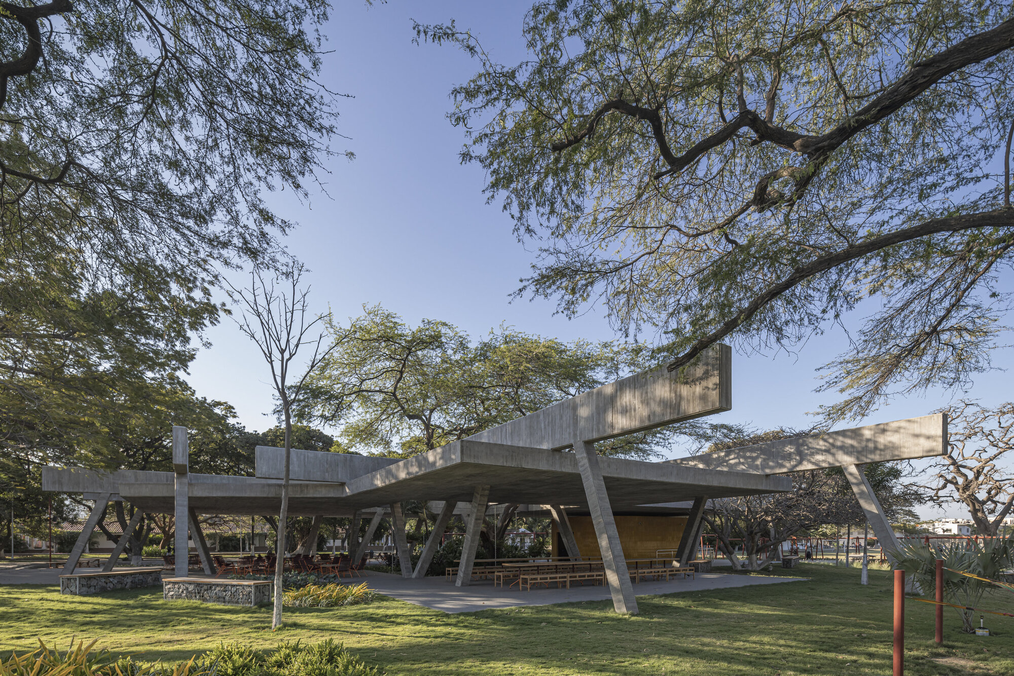 Wide view of the concrete canopy on slanted columns, set on a lawn beneath spreading tree branches and blue sky