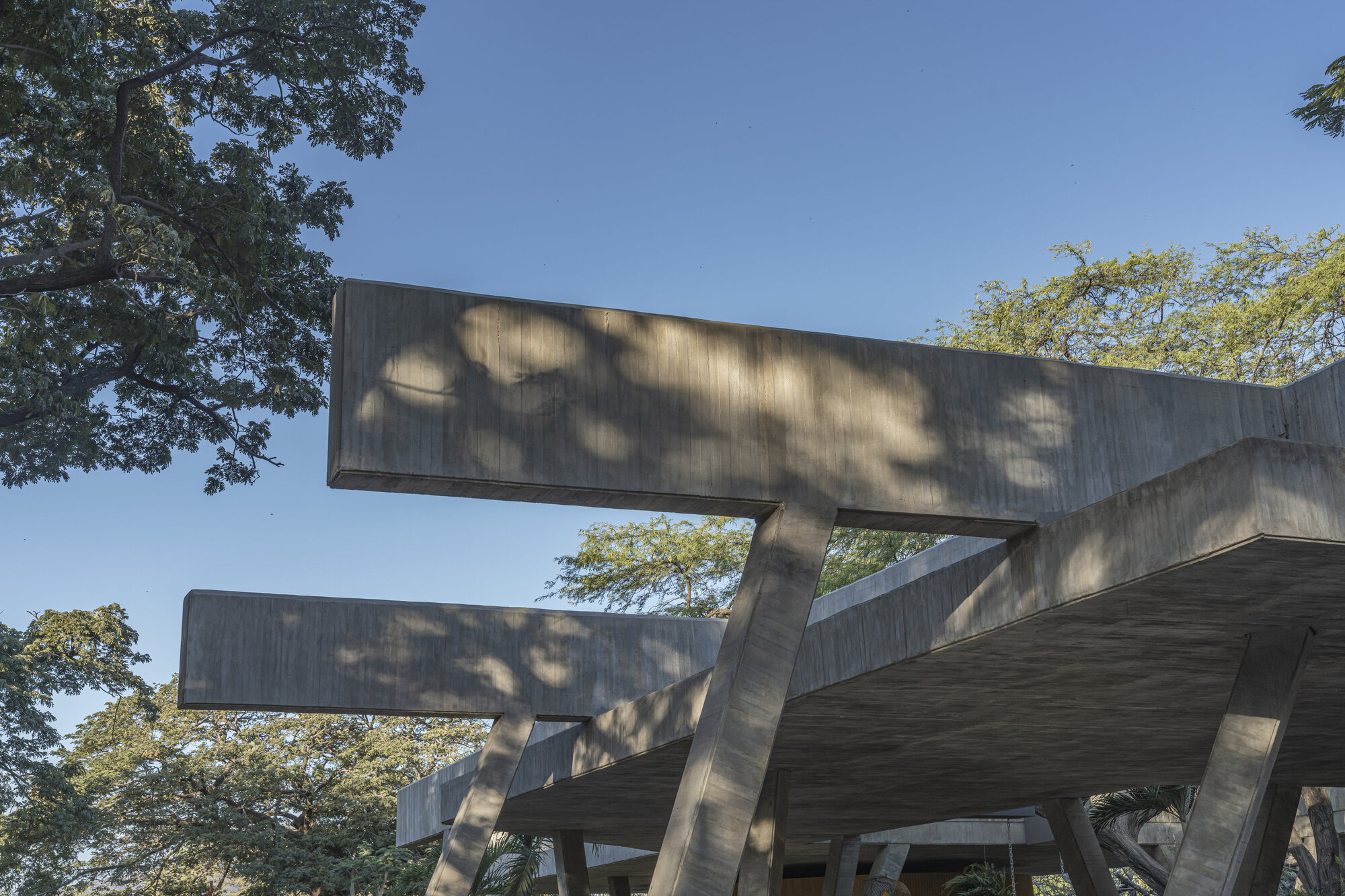 Upward view of cantilevered concrete beams and angled columns, with dappled leaf shadows on the surface under a clear blue sky