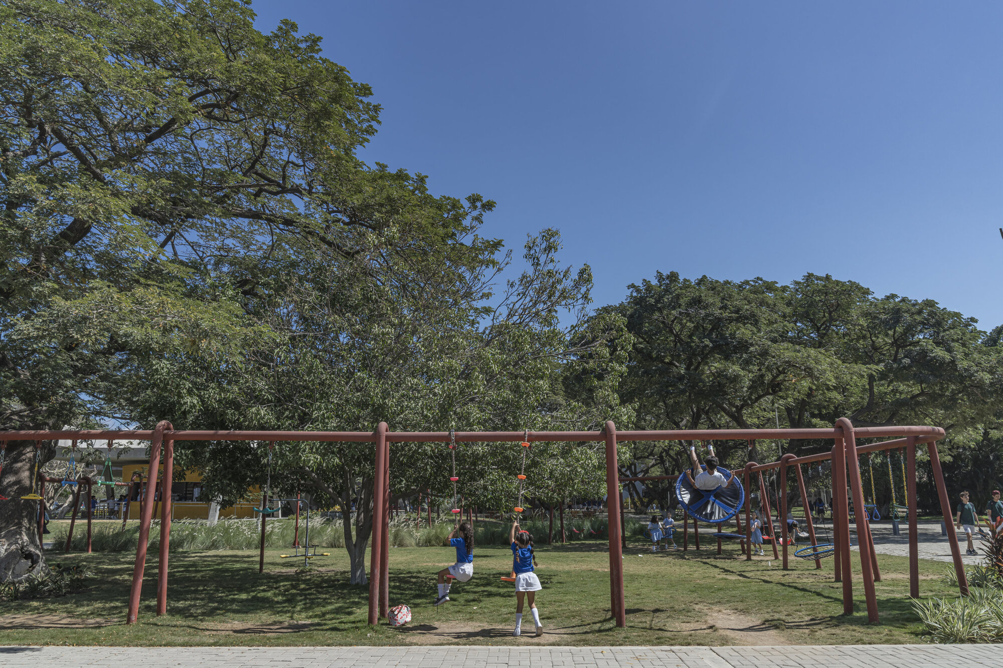 Children in blue uniforms on rust-red steel swing frames in a shaded park lawn under large trees and a deep blue sky