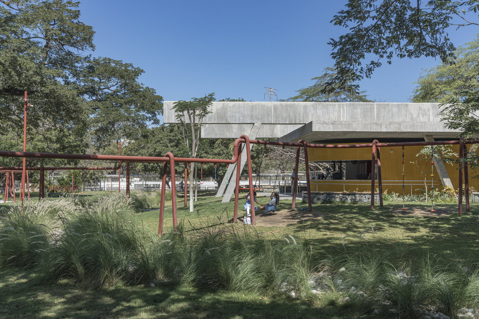Concrete roof slab over a yellow-clad pavilion seen past red steel play frames and clumps of ornamental grass in strong afternoon sun