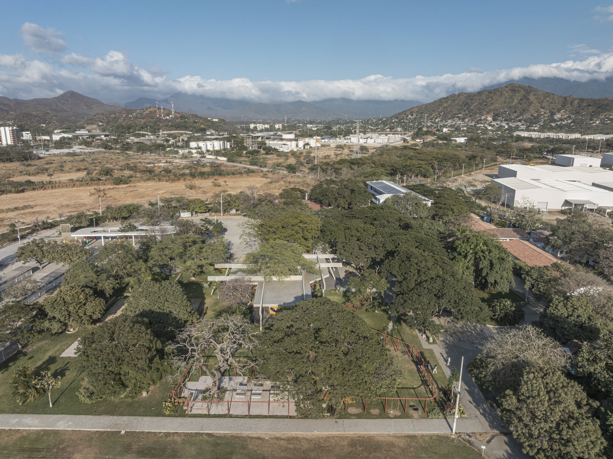 Aerial view of the low concrete pavilion among mature trees and a red play structure, with hills and cloud beyond
