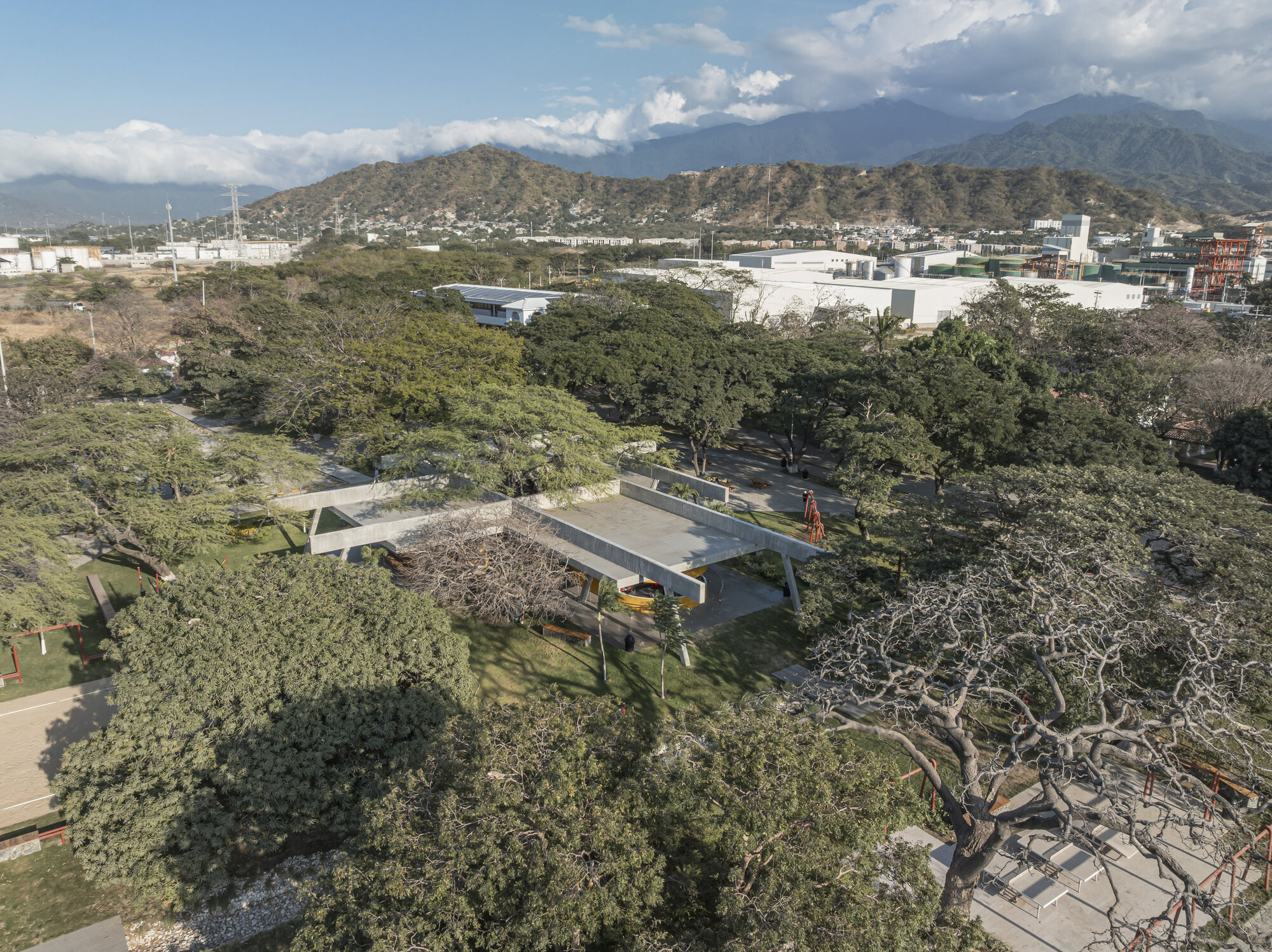 Oblique aerial view of the concrete roof with angled supports beneath a leafy canopy, with a mountain ridge behind under scattered cloud