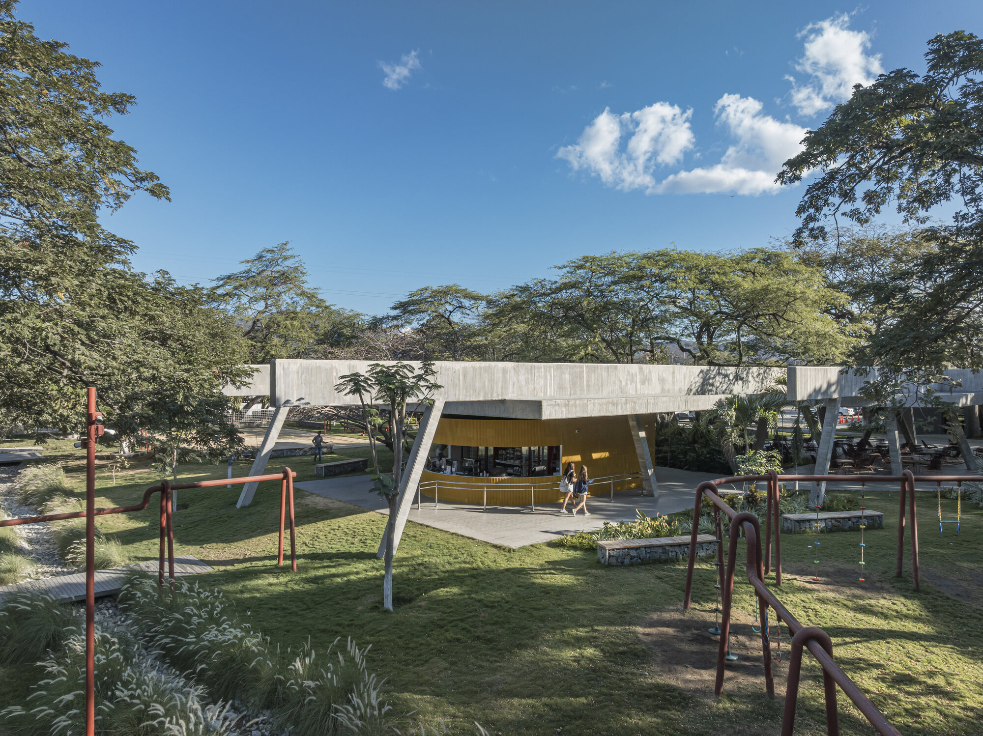 Concrete canopy on slanted columns over a yellow-walled kiosk, seen across lawn and red play bars under a blue sky