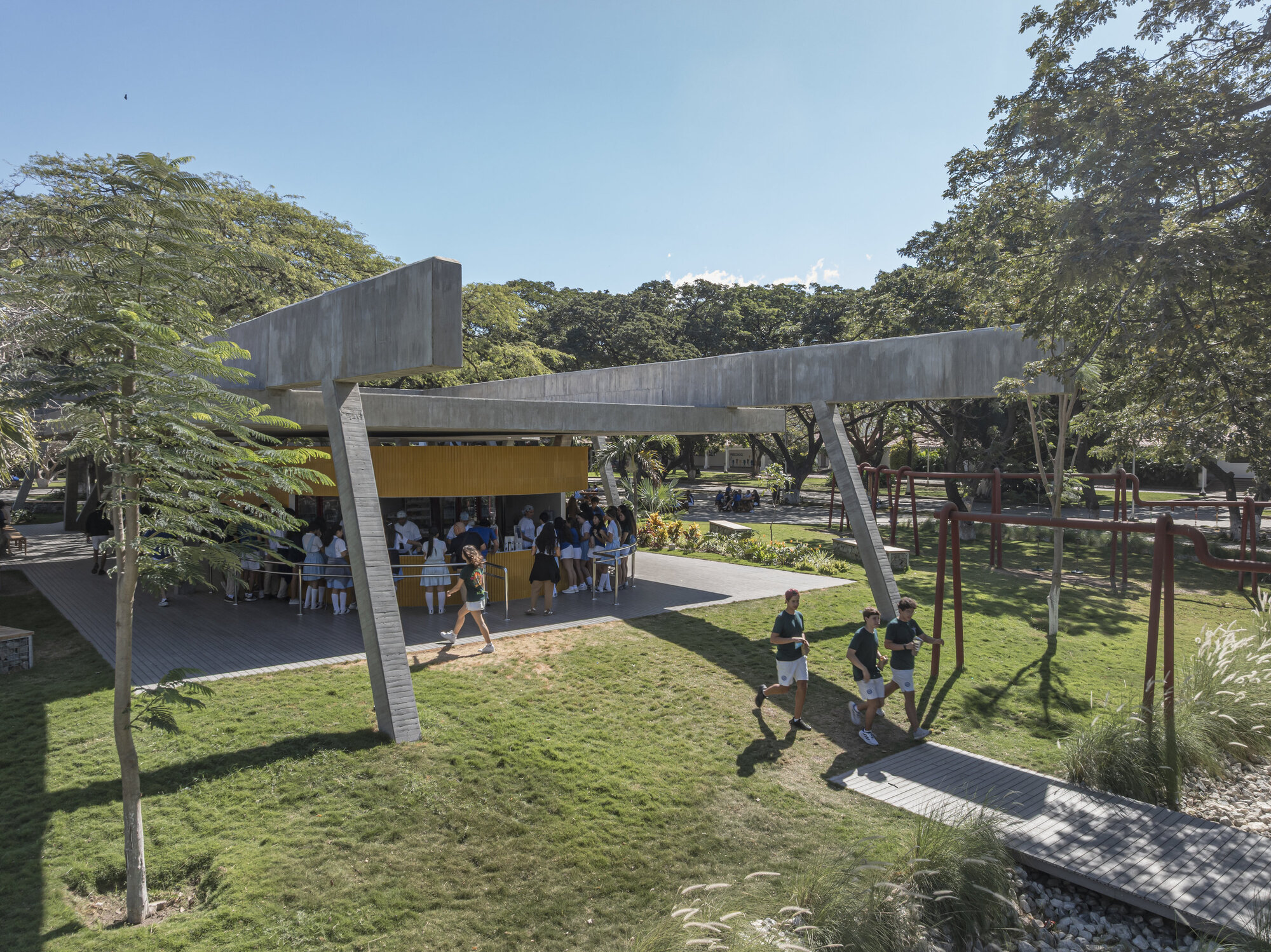 Students gathered at the yellow kiosk under a angled concrete canopy while three boys walk across the sunlit lawn