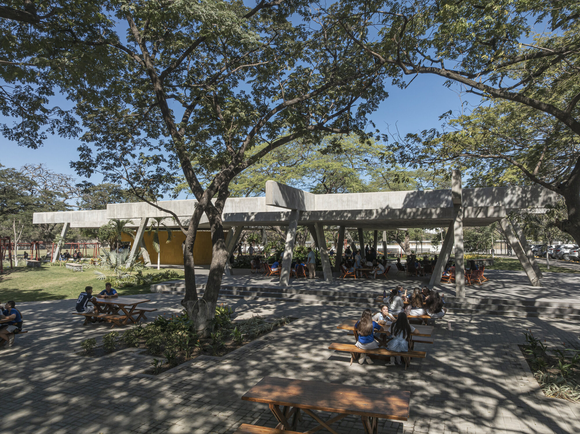 Flat concrete canopy on slanted columns above picnic tables, with dappled shade from large trees and students eating lunch