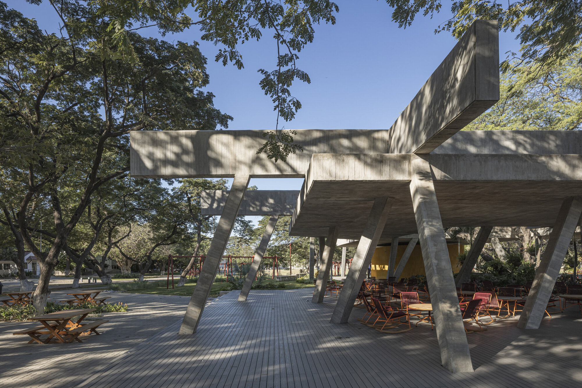 Angled concrete columns and cantilevered beams over a timber deck with red chairs, under strong afternoon sun and tree shadows